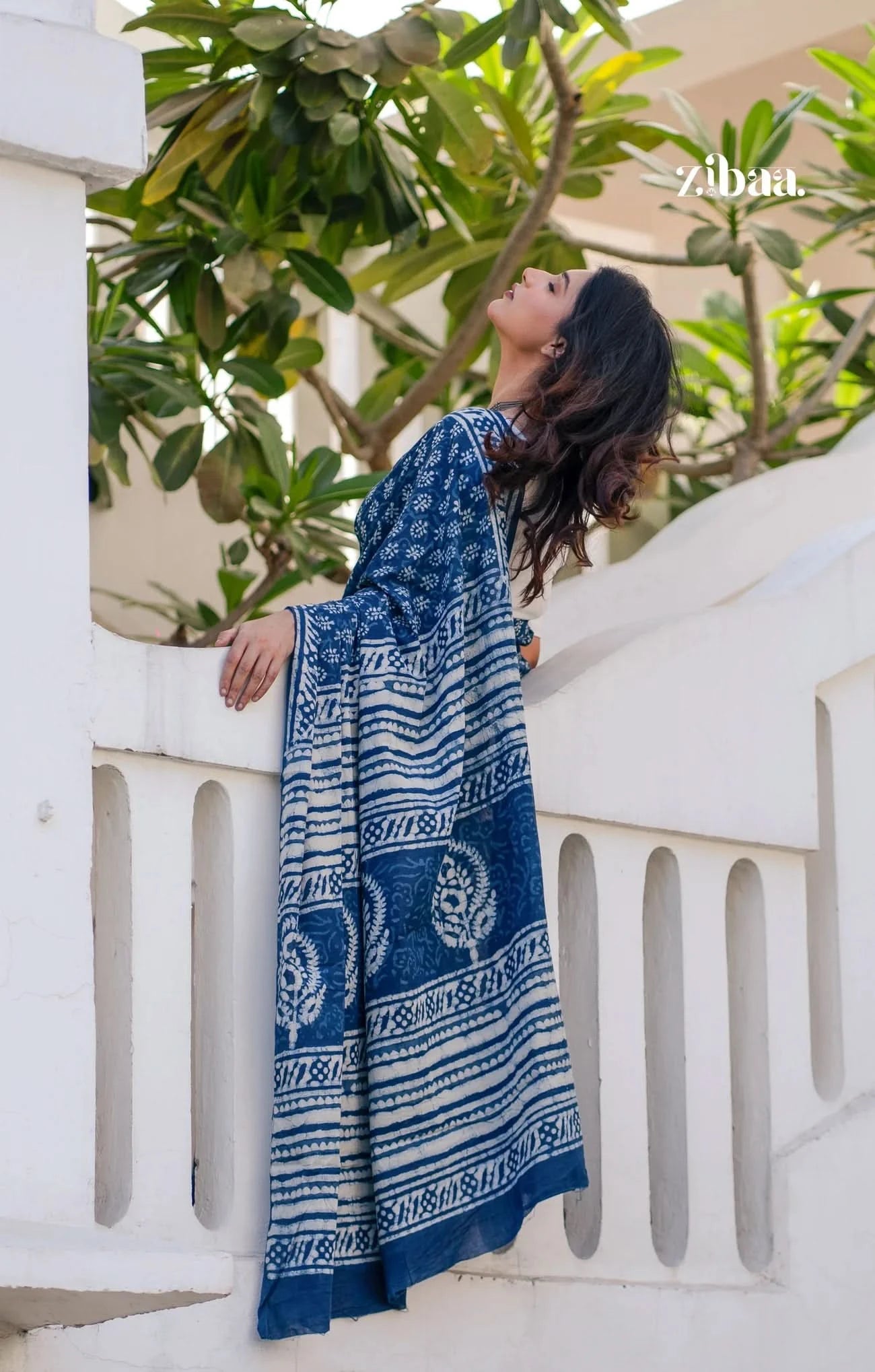Woman wearing a blue patterned dress standing on a white railing with greenery in the background