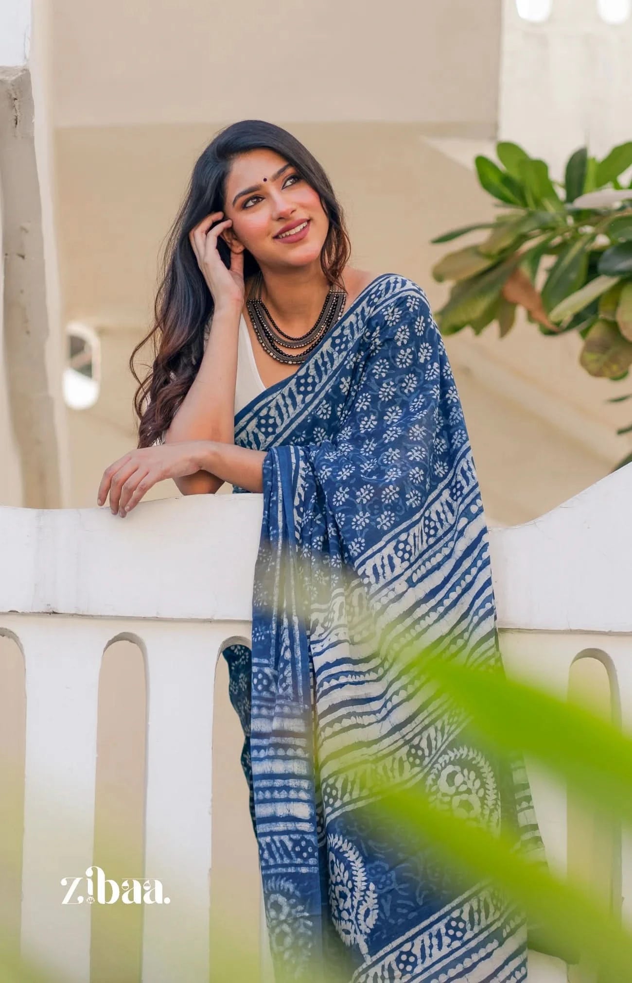 Woman wearing a blue and white patterned saree leaning on a railing with greenery in the background