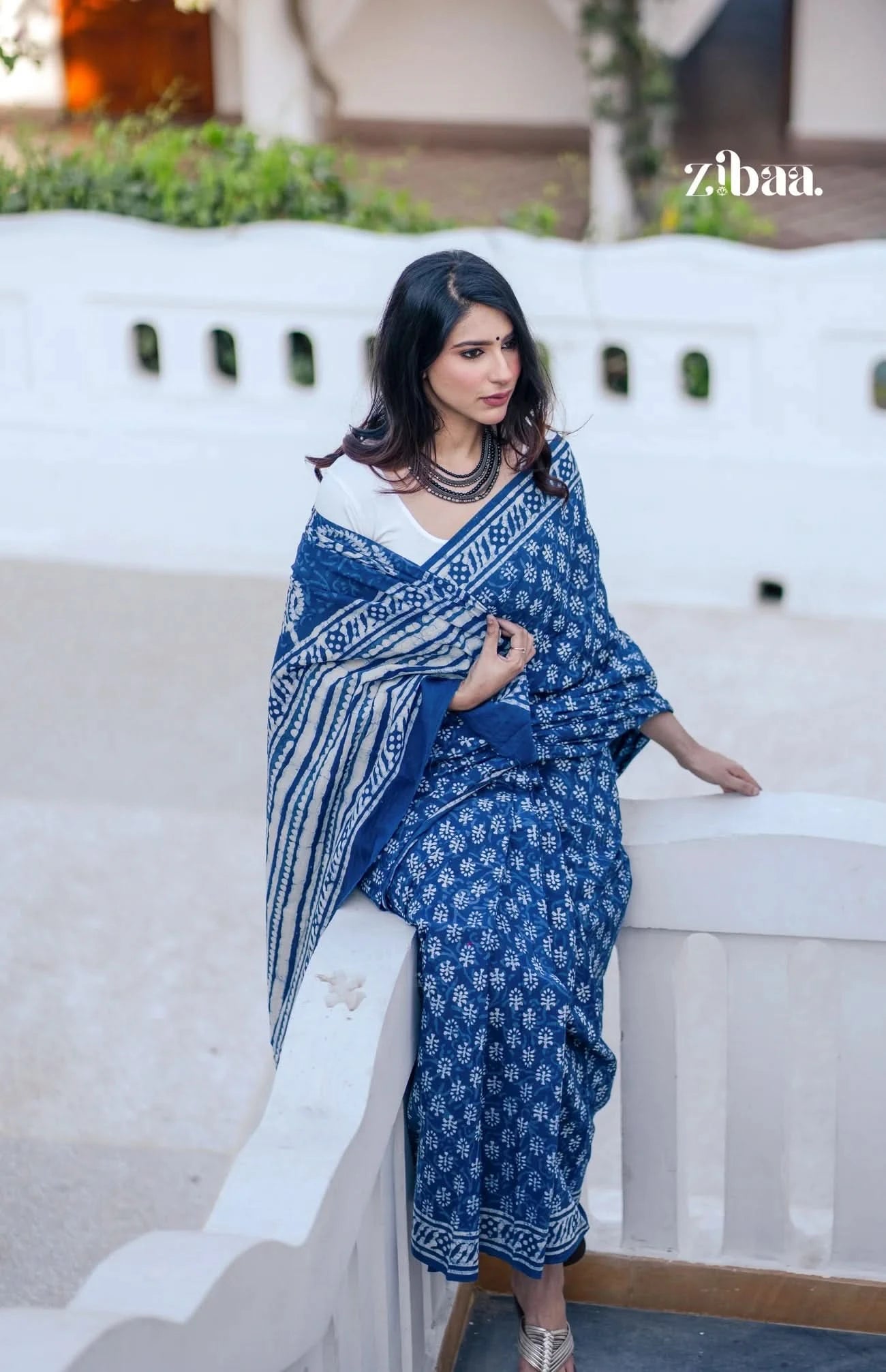 Woman in a blue and white patterned saree sitting on a white bench with 'zibaa' branding.