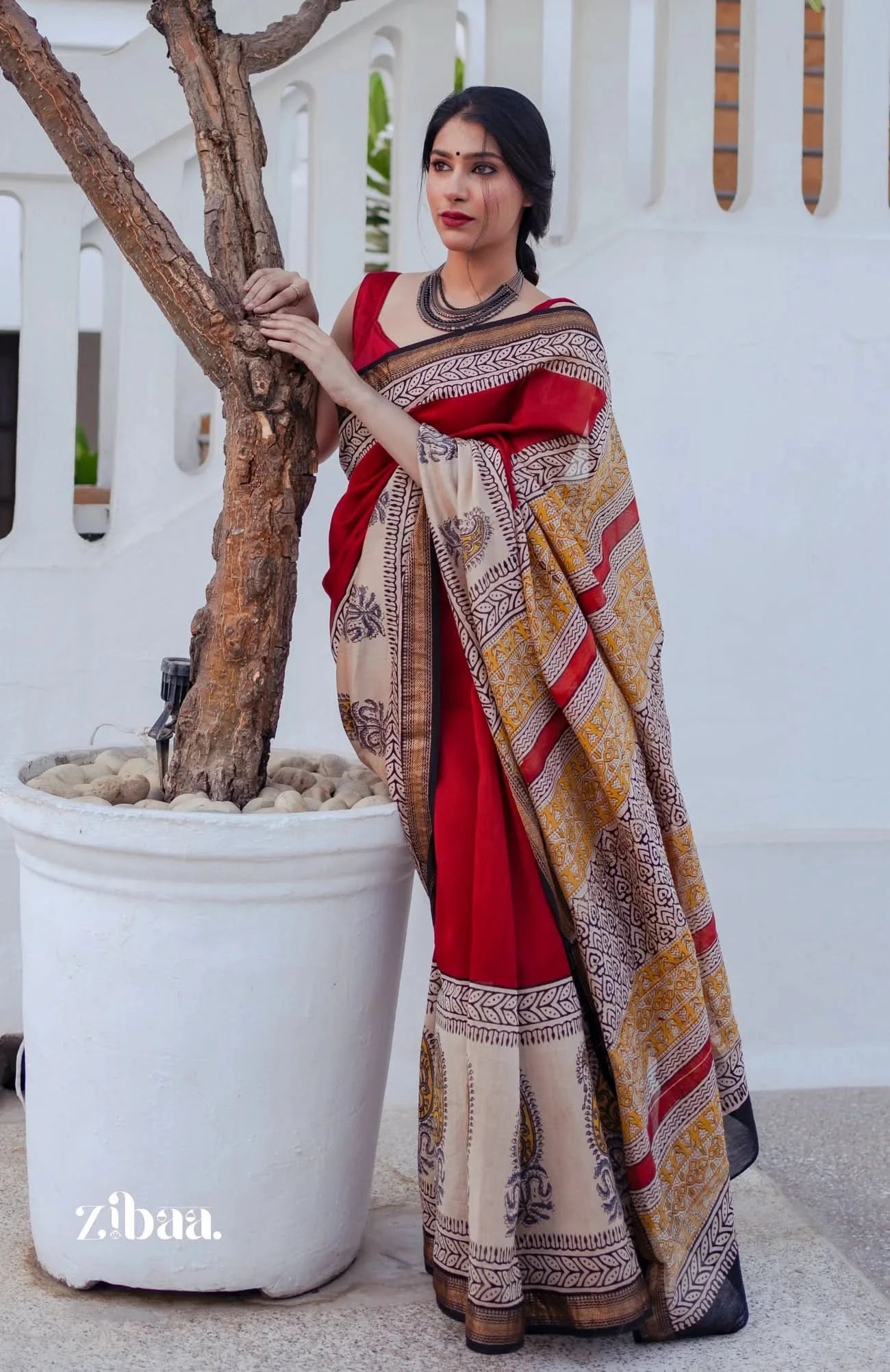 Woman in a red and patterned saree standing next to a white pot with a tree