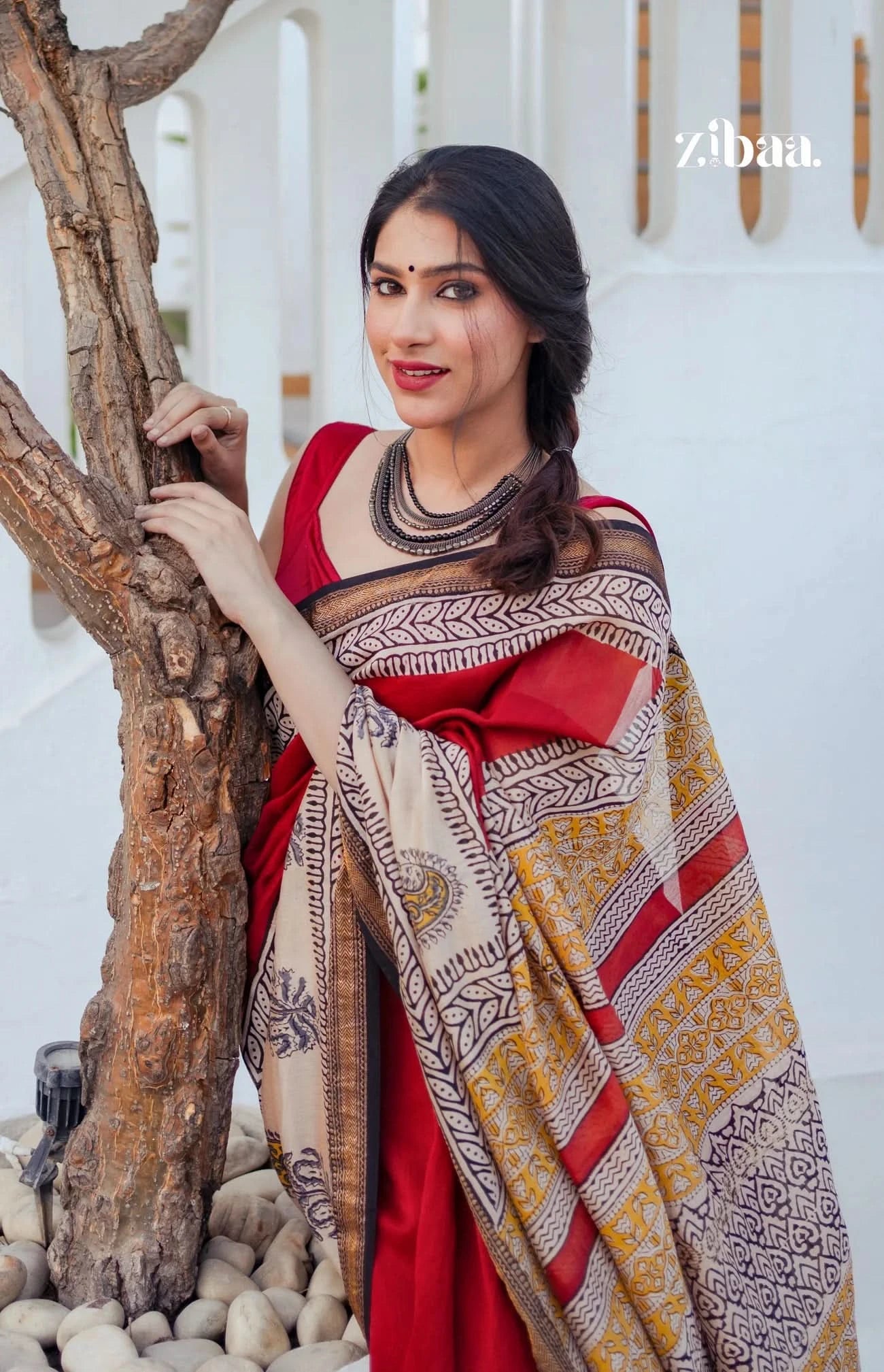 Woman wearing a patterned shawl over a red dress, standing against a white wall with 'zibaa' branding.