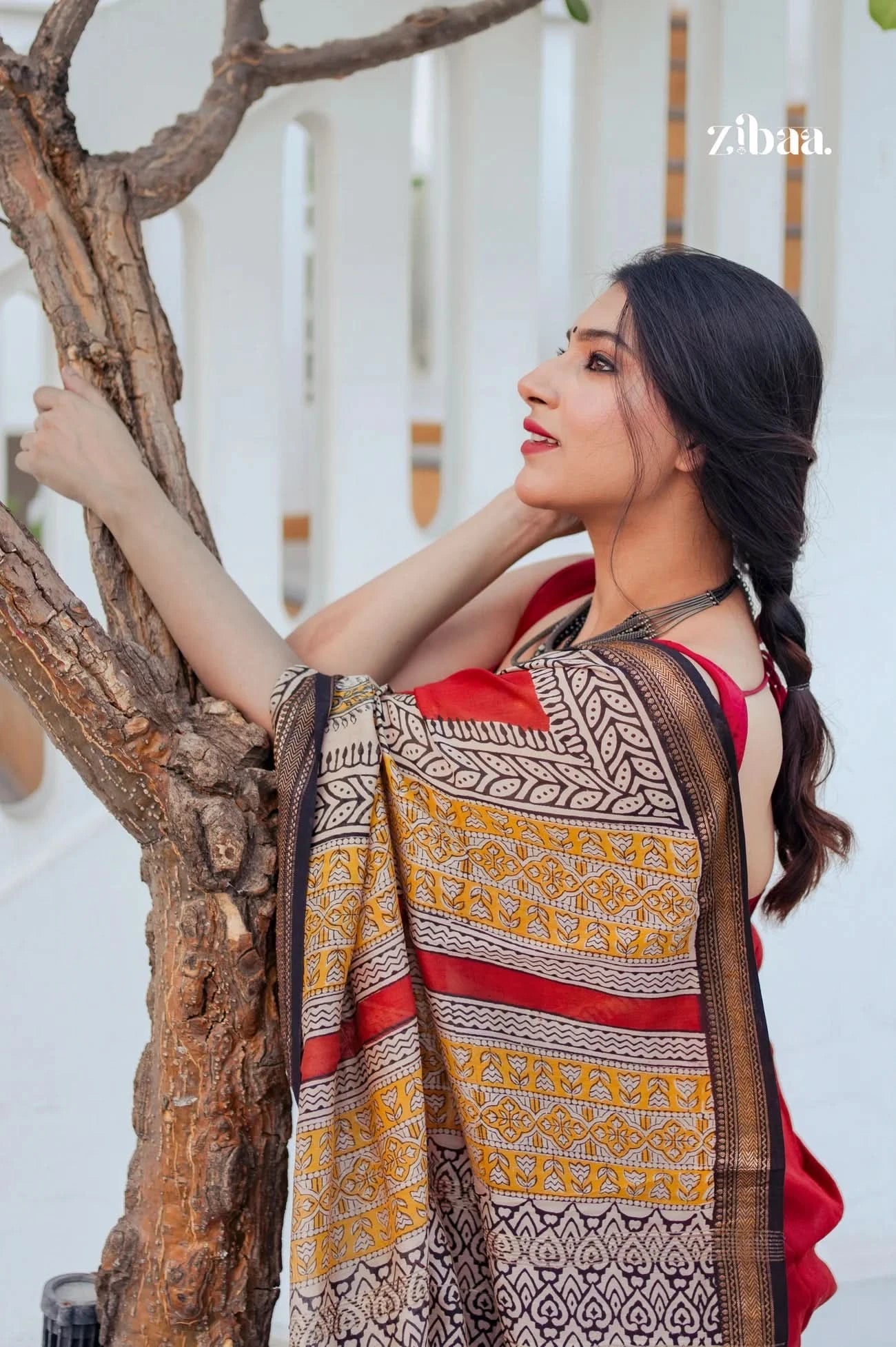 Woman wearing a patterned dress with a red top, leaning against a tree with a white background.
