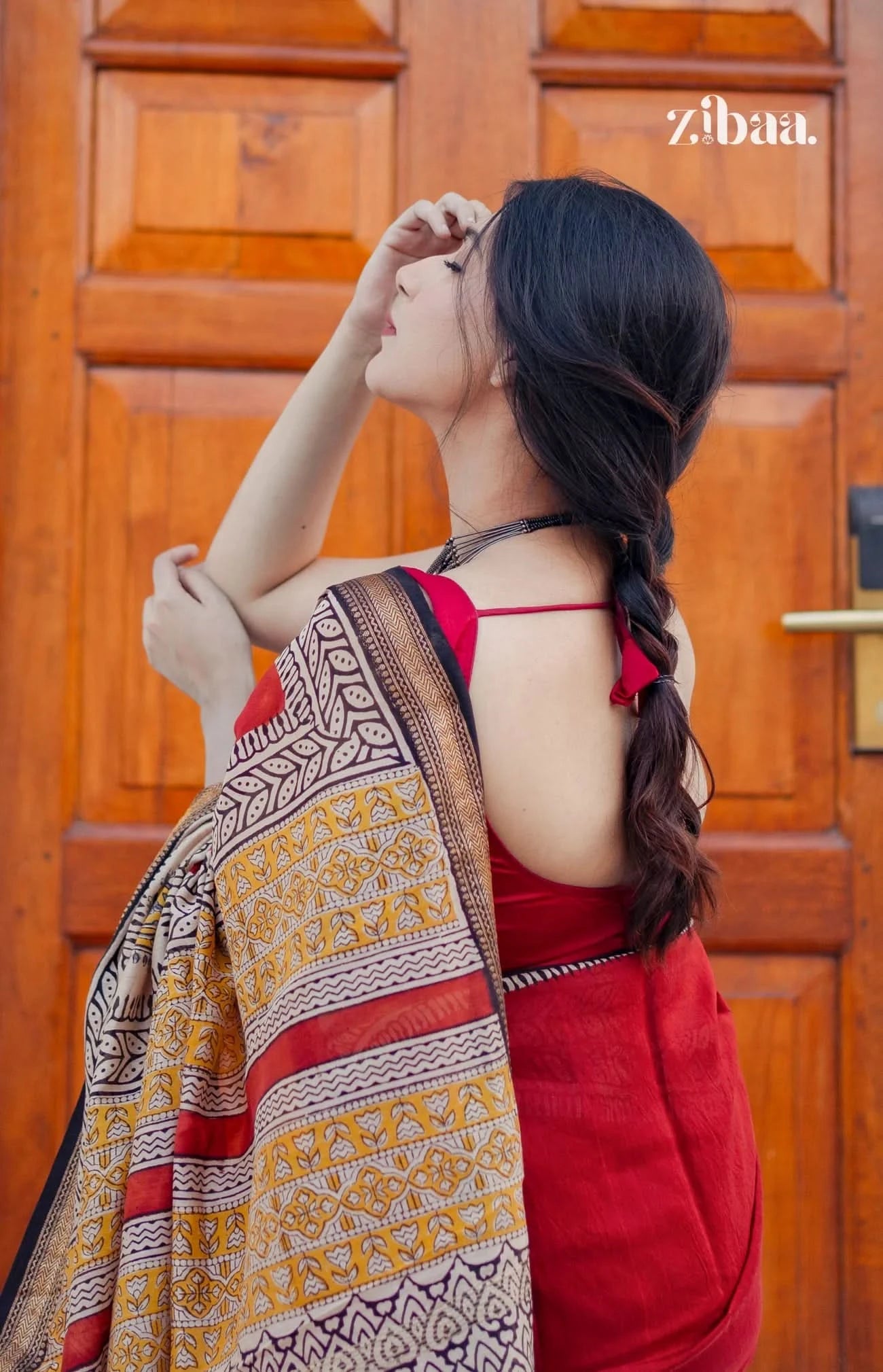 Woman in a red saree with a patterned shawl standing in front of a wooden door, with 'zibaa' branding.