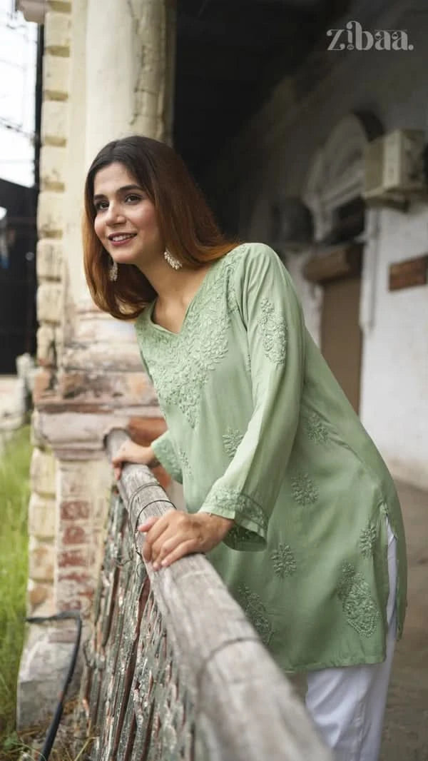 Woman in a light green embroidered top and white pants standing on a balcony.