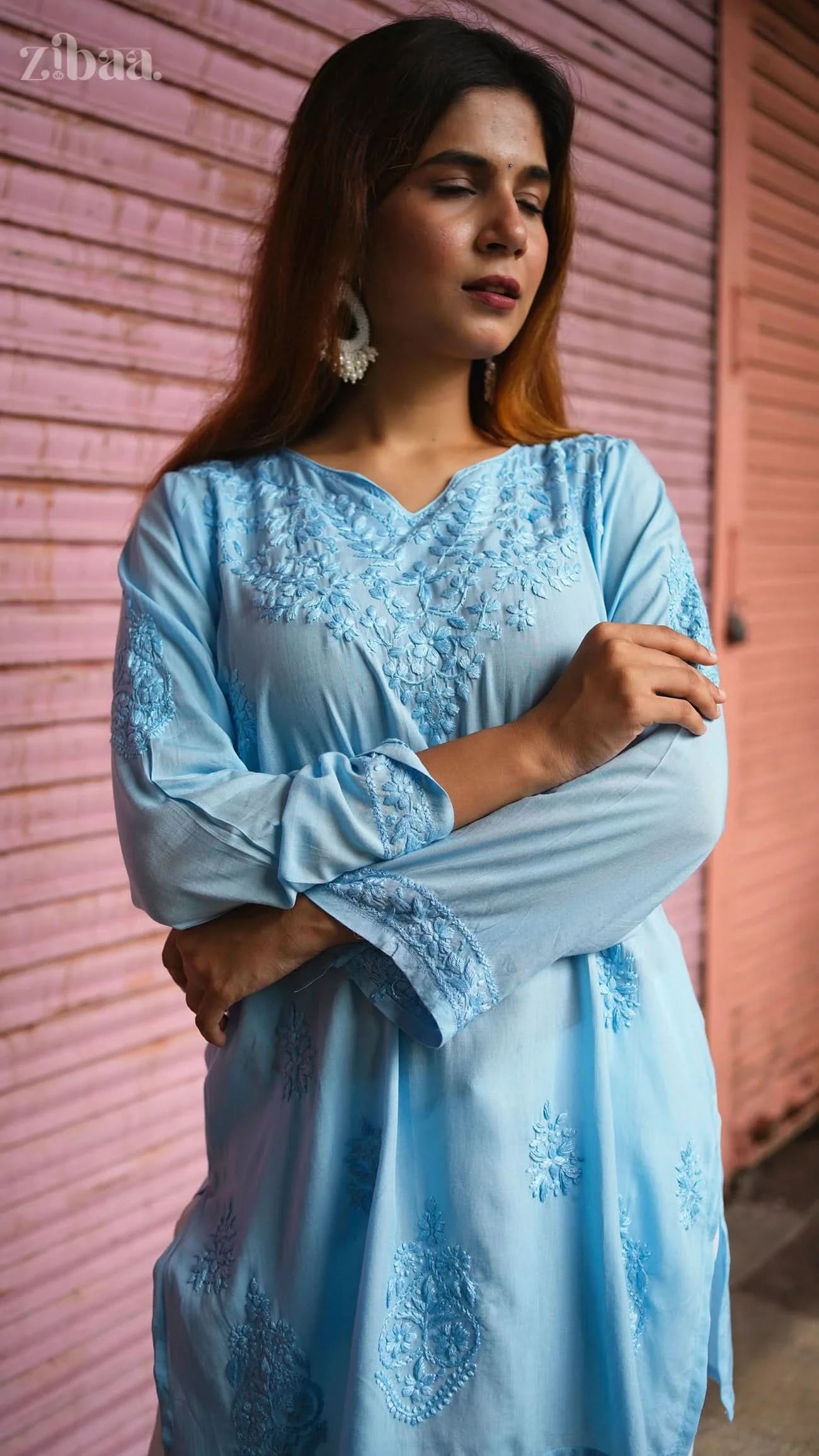 Woman wearing a light blue embroidered dress against a pink wall.