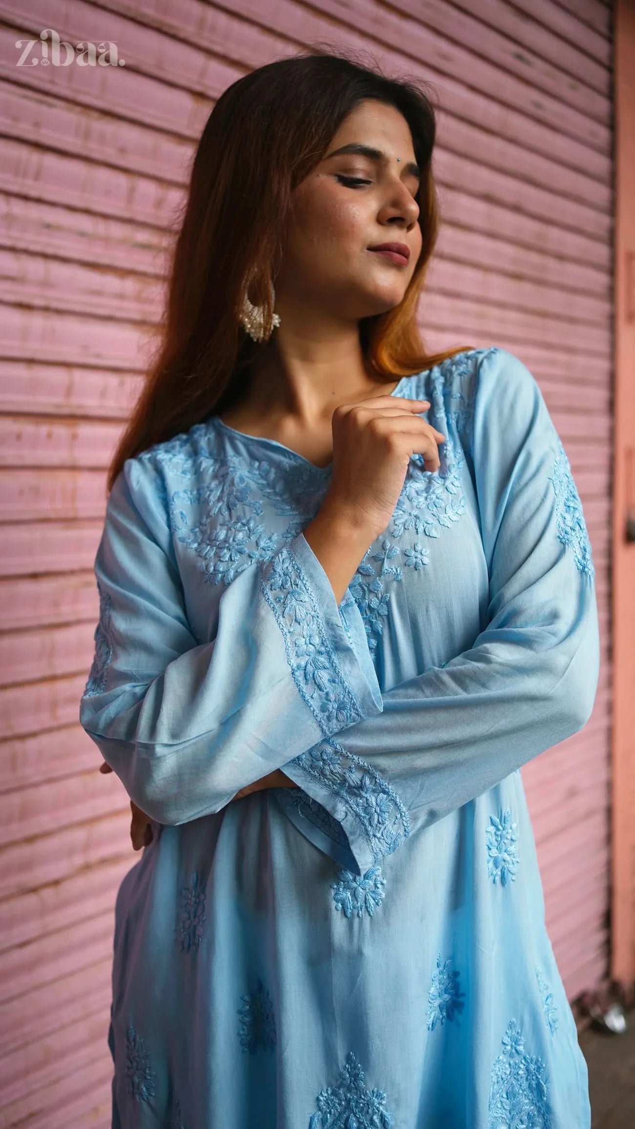 Woman wearing a light blue embroidered dress against a pink wall.