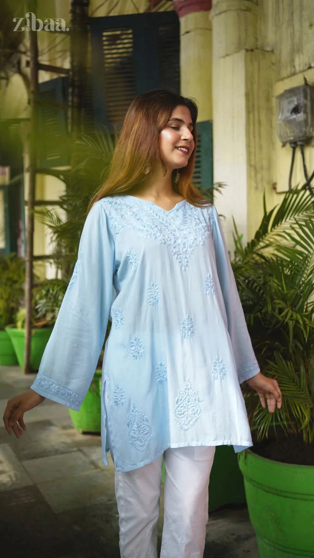 Woman wearing a light blue embroidered top and white pants standing outdoors with green plants in the background.