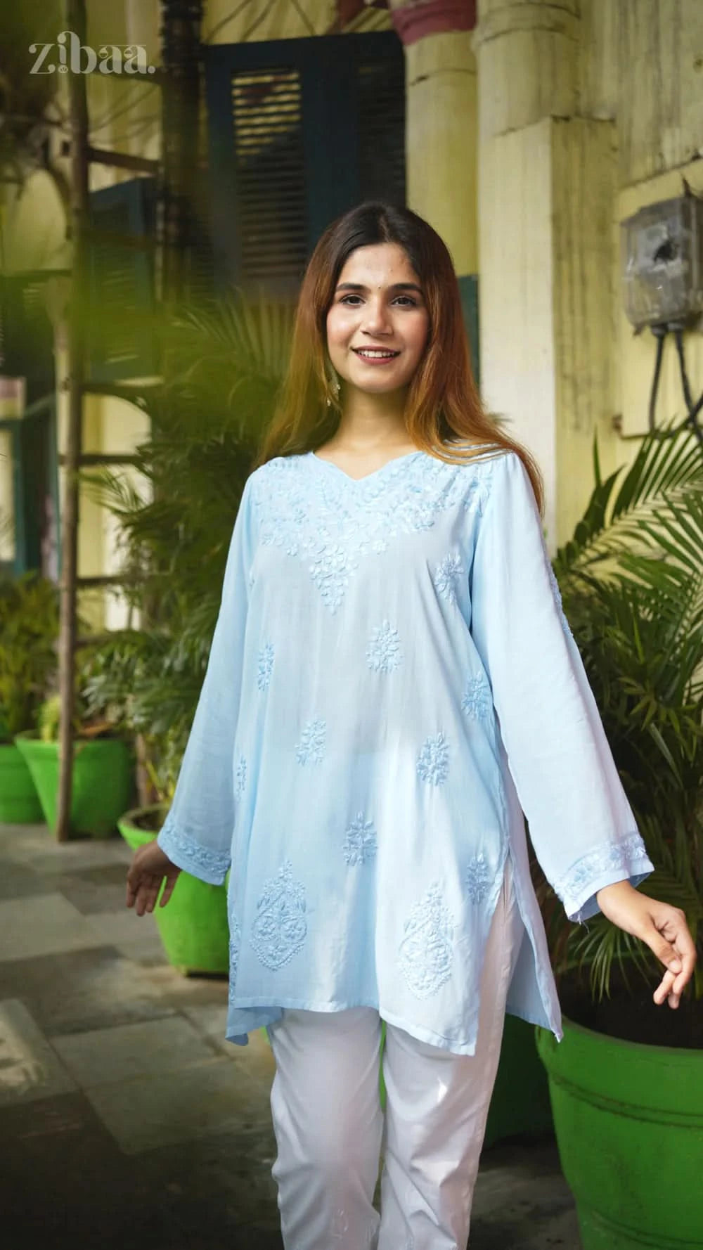 Woman wearing a light blue embroidered top and white pants standing outdoors.