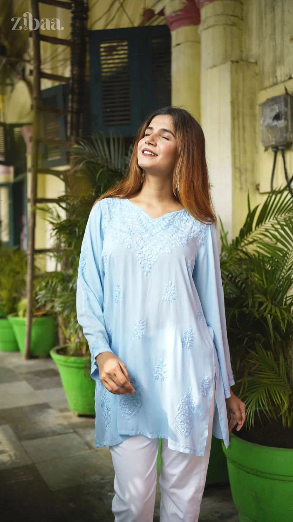 Woman wearing a light blue embroidered top and white pants standing outdoors with plants in the background