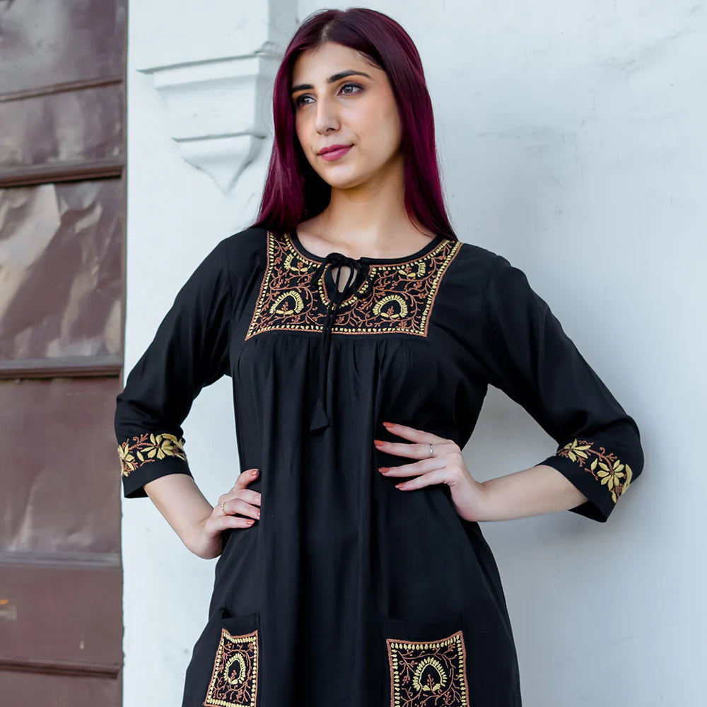 Woman wearing a black dress with gold embroidery against a white wall.