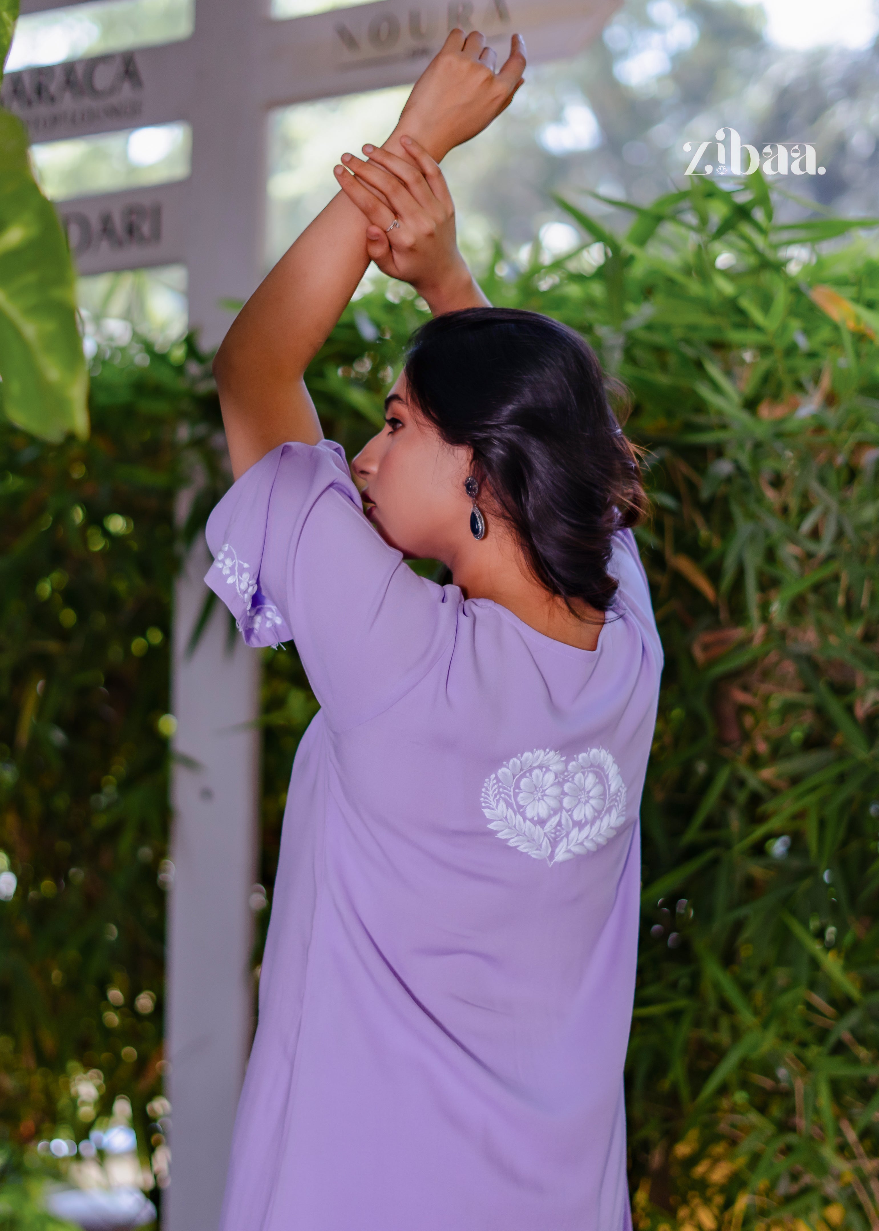 The model showcases the Husn Lavender Luxury Rayon Chikankari Set with Elegant Back Design while posing outdoors. Husn Lavender Luxury Rayon Chikankari Set with Elegant Back Design features intricate white embroidery that stands out on lavender rayon. The graceful pose highlights its festive and premium appeal.