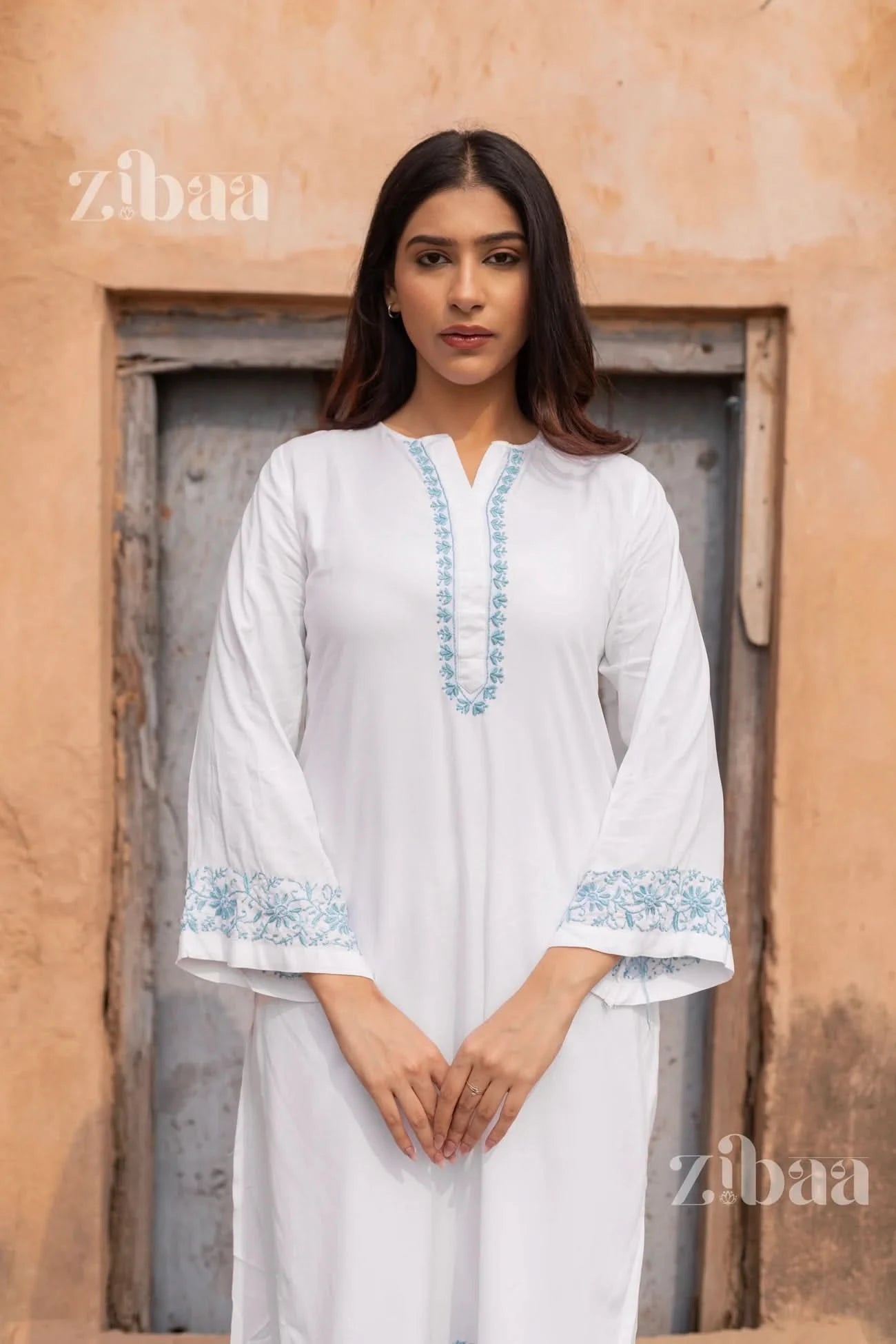 Woman wearing a white outfit with blue embroidery in front of a rustic door, with 'zibaa' branding.