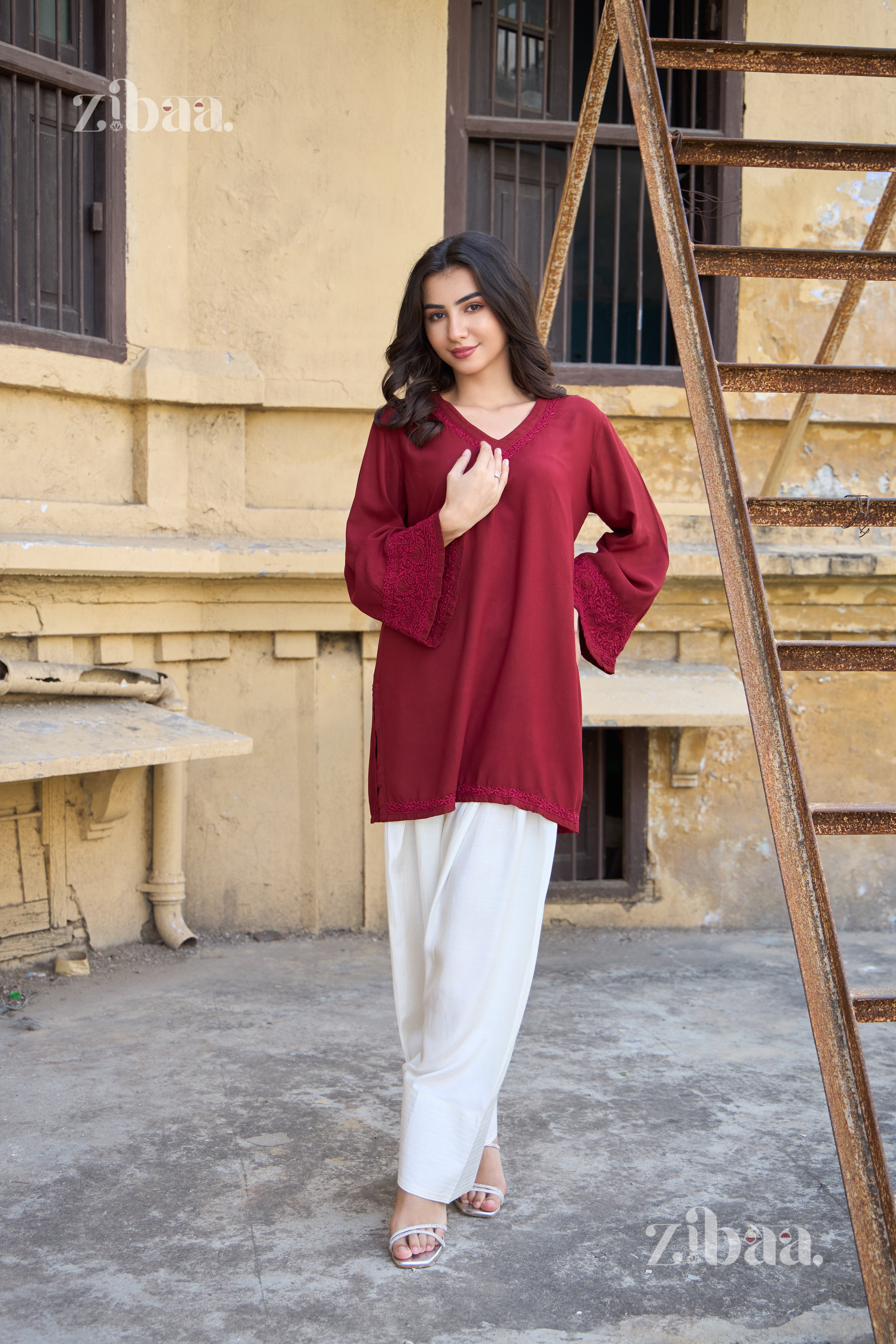 Woman in a red top and white pants standing in front of a textured wall with a ladder.