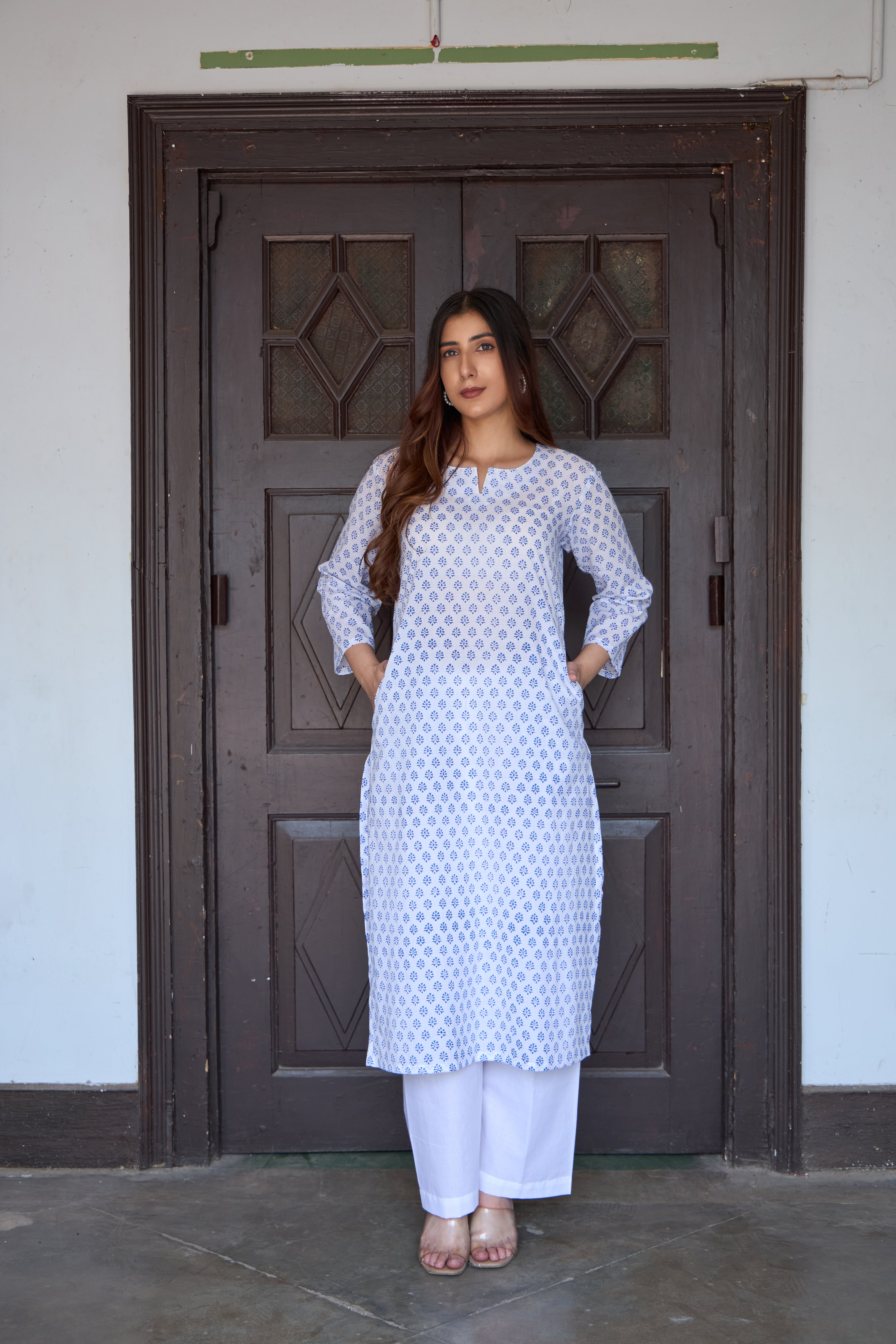 Woman wearing a white and blue patterned kurta standing in front of a dark wooden door.