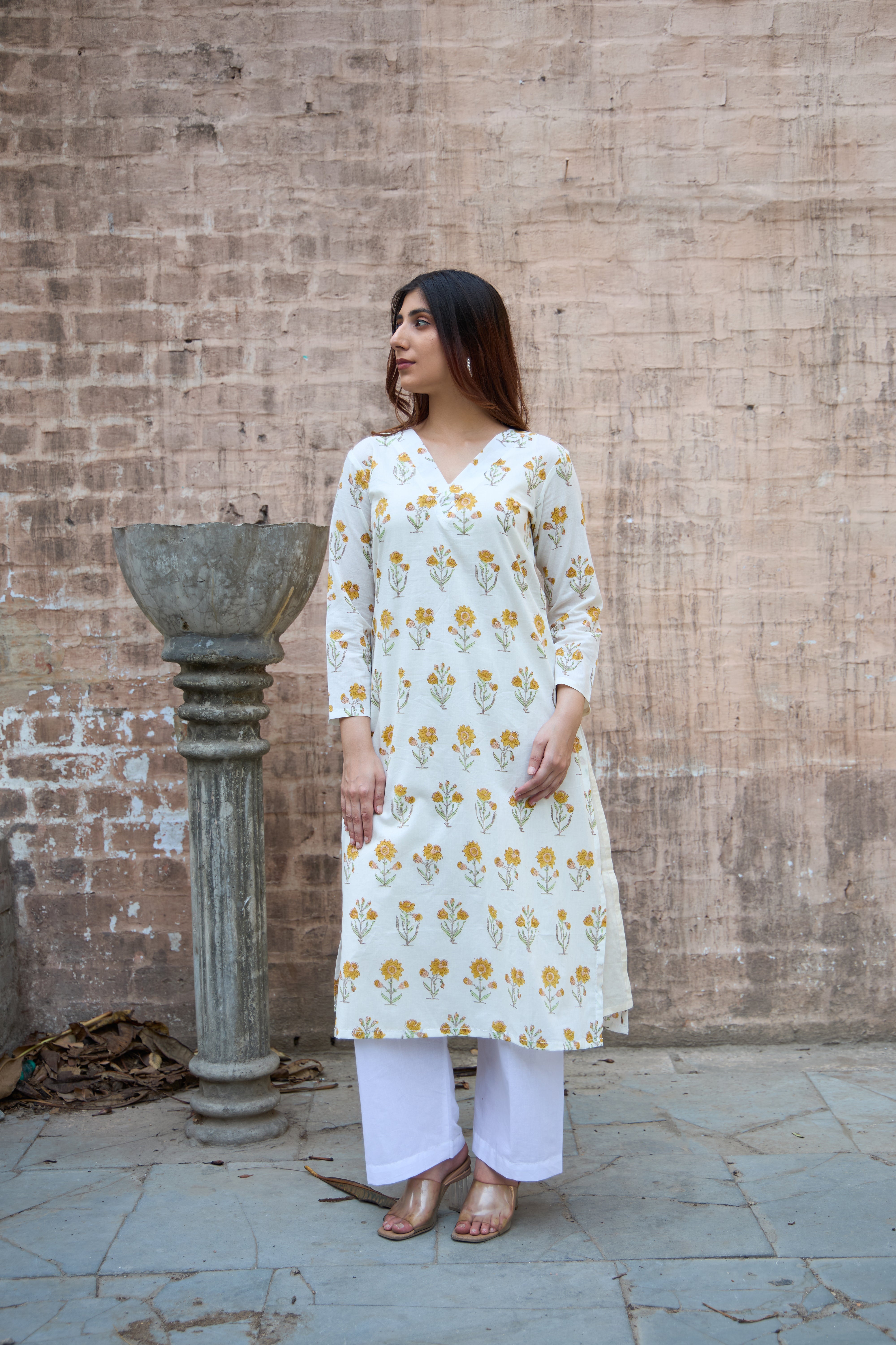 Woman wearing a white kurta with yellow floral patterns against a brick wall.