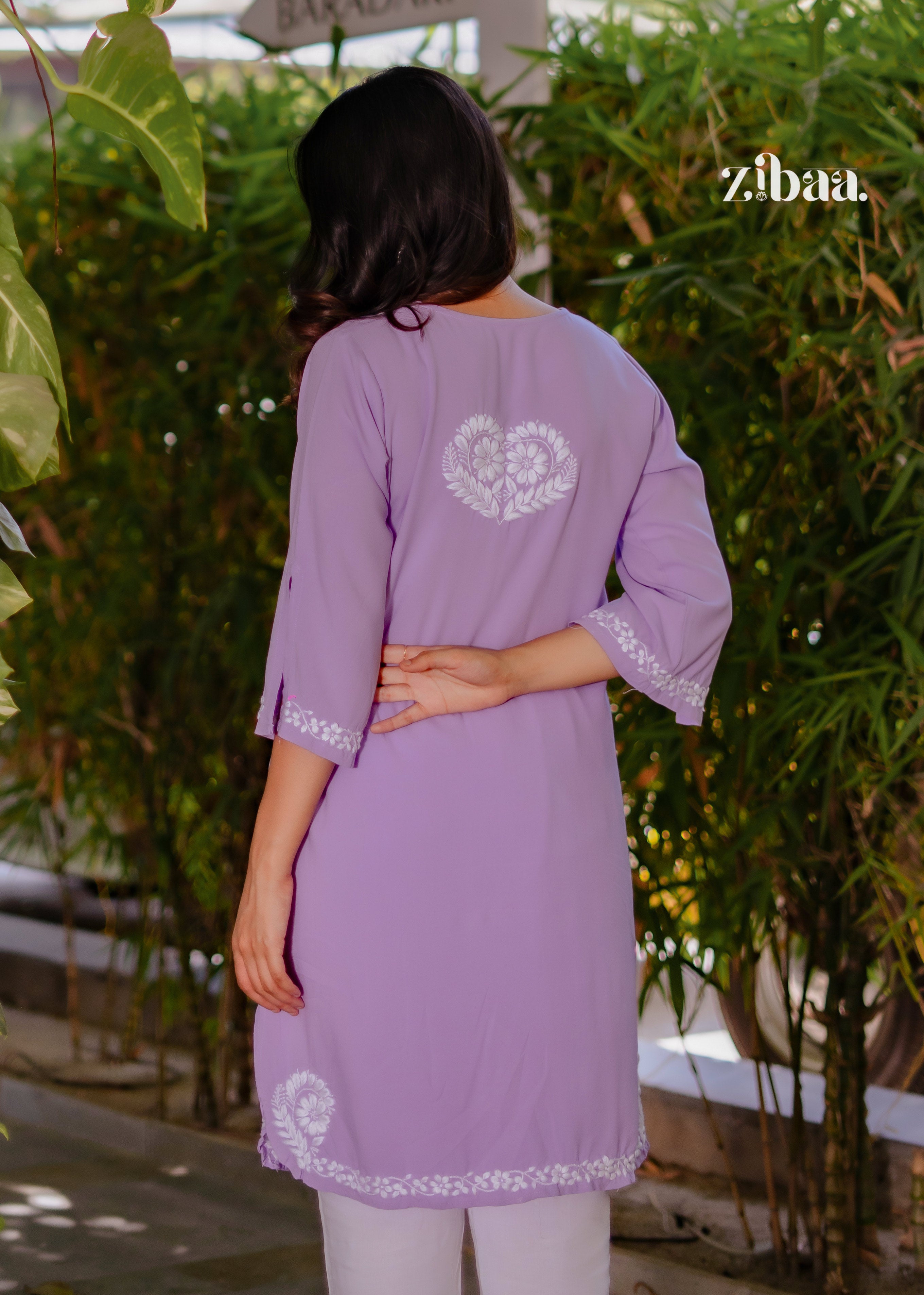 he model poses gracefully from the back in a Husn Lavender Luxury Rayon Chikankari Co-ord Set with Back Embroidery. Husn Lavender Luxury Rayon Chikankari Co-ord Set features intricate white floral motifs on soft lavender rayon. The elegant embroidery highlights its premium craftsmanship.