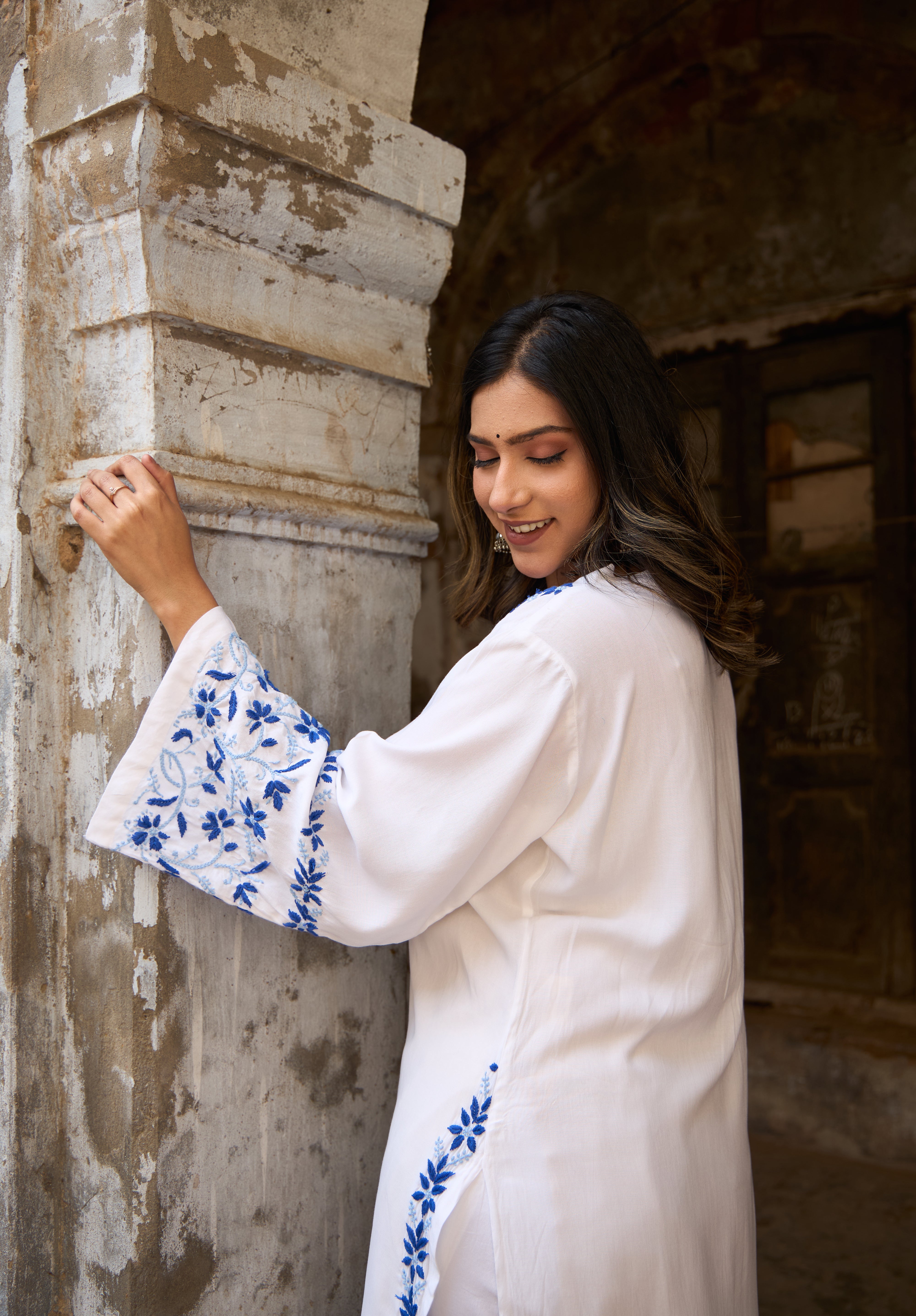 Woman in a White Chikankari Kurta with blue embroidery standing against a stone wall.