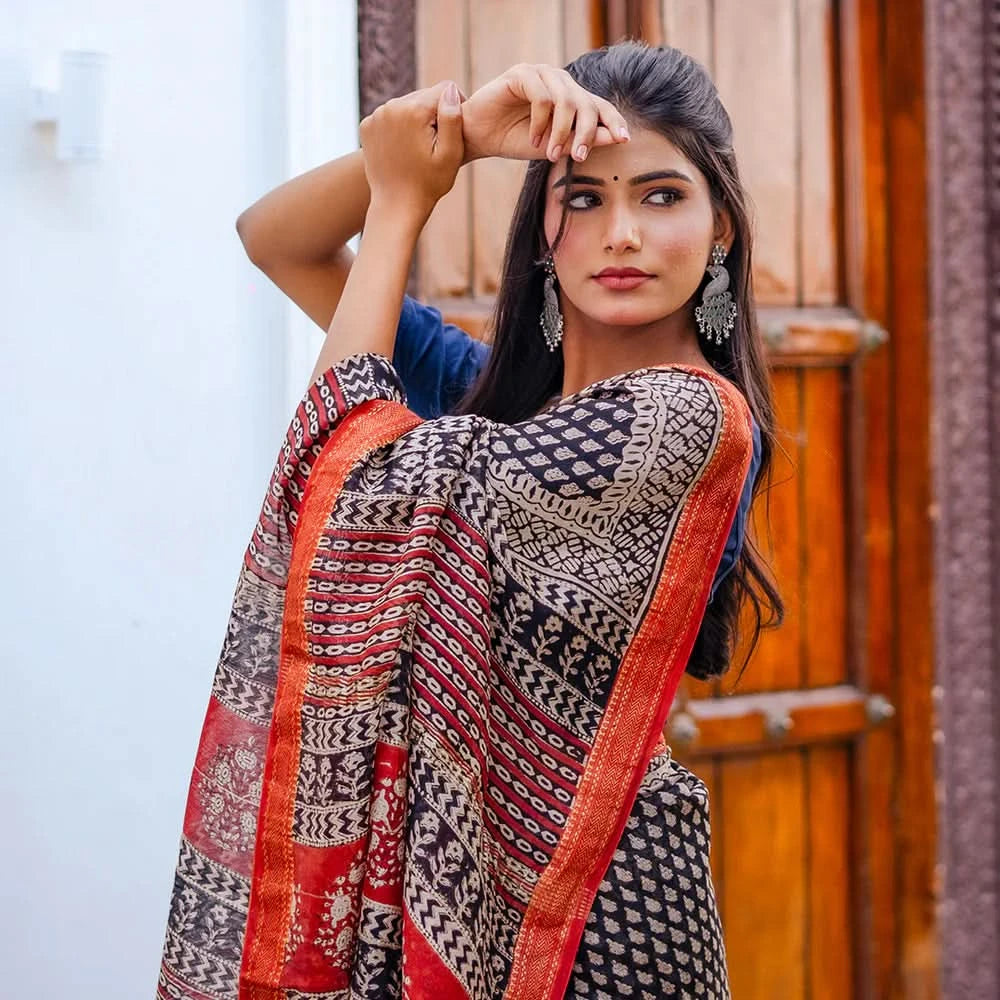 Woman wearing a traditional patterned saree with a red border, standing against a wooden door.