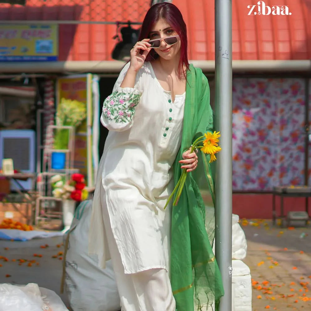 Woman in a white outfit with floral embroidery holding a green scarf and yellow flowers, standing against a colorful background.
