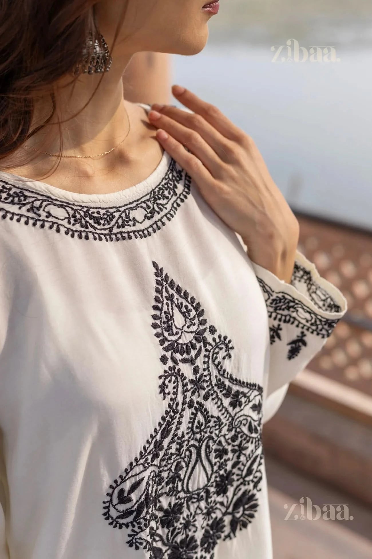 Close-up of a woman wearing a white Chikankari kurta set with intricate black embroidery, gently touching the neckline, standing outdoors.