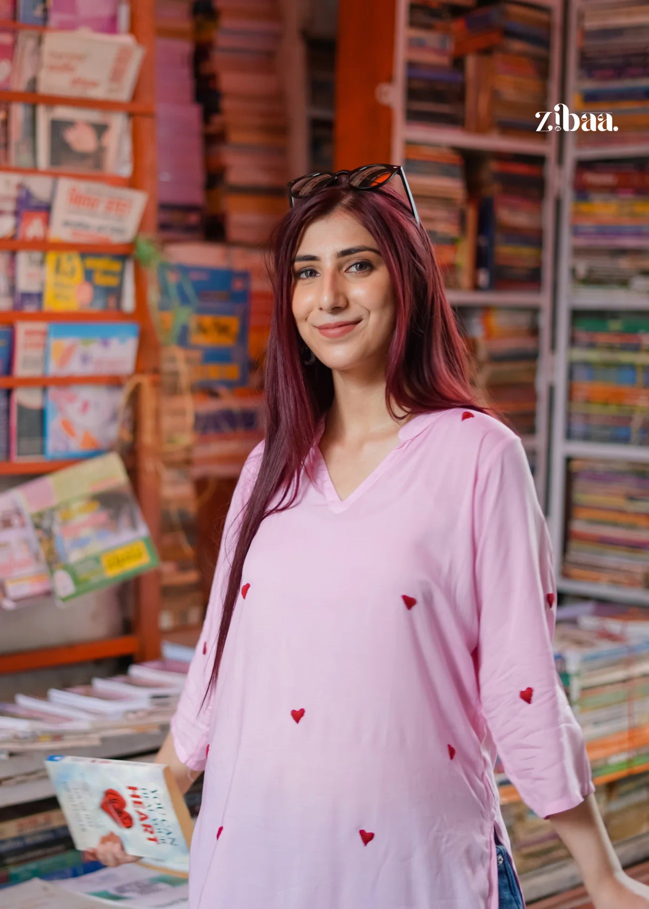 The model stands on a street, wearing a pink chikankari kurti with heart motifs, with a book stall in the background.