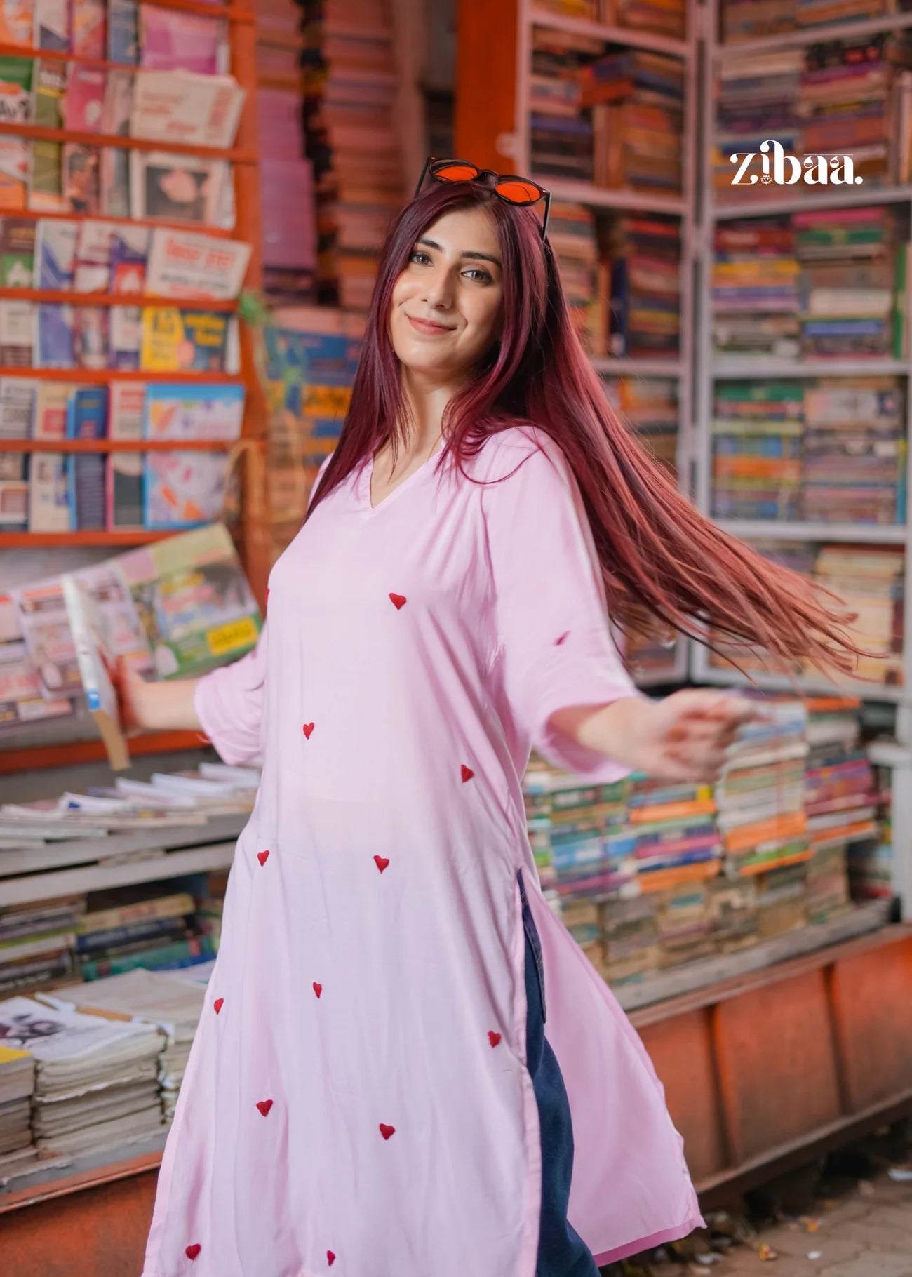 The model is slightly twirling on a street, wearing a pink chikankari kurti with heart motifs, with a book stall in the background.