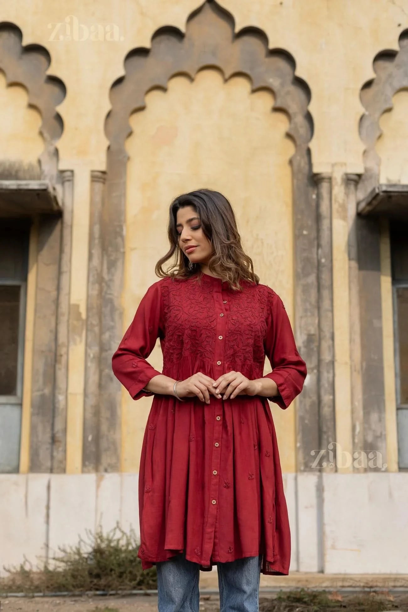 A woman stands outdoors with her hands gently clasped, wearing a maroon Chikankari top, highlighting its intricate embroidery.