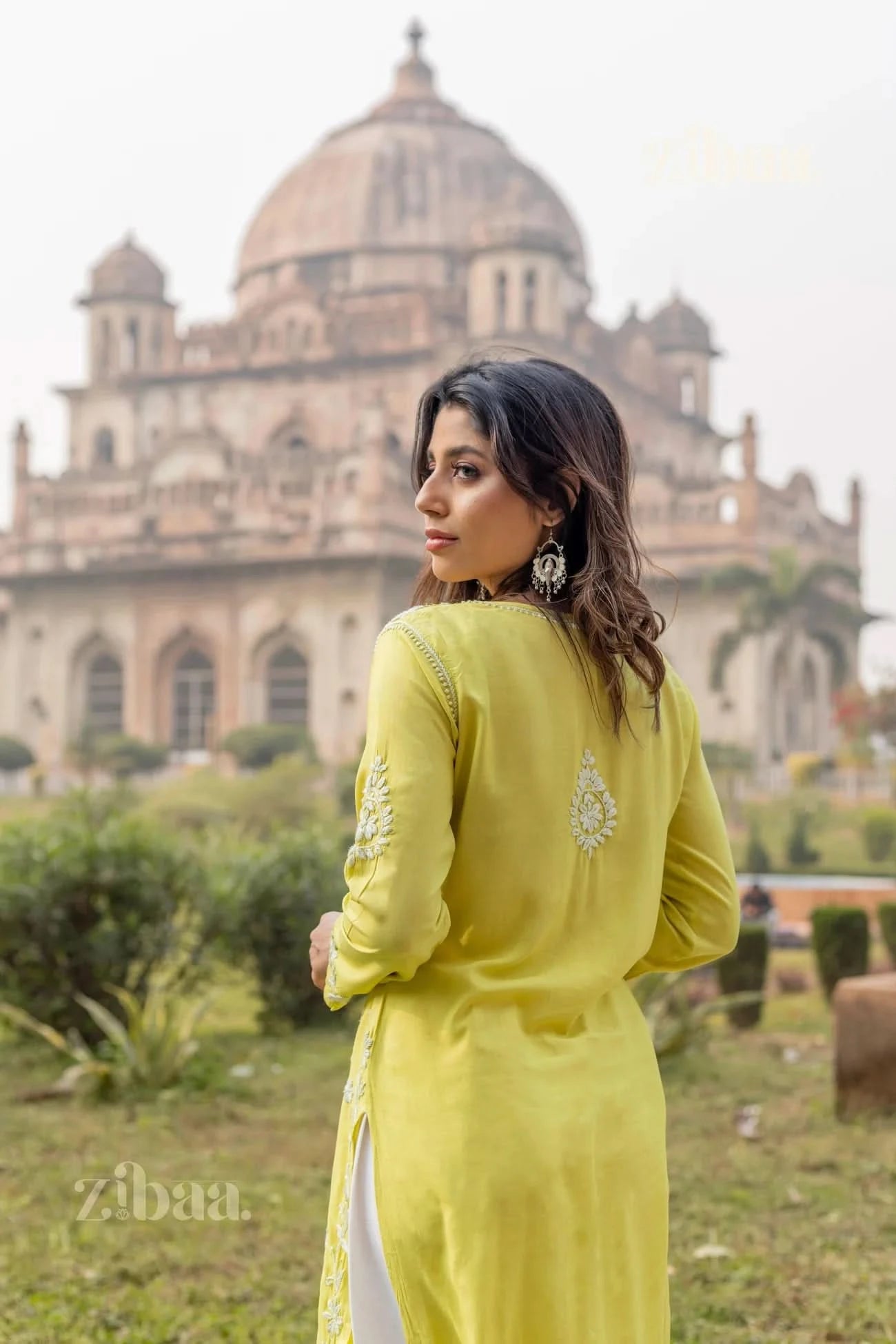 A woman stands in an outdoor garden, facing slightly away, showcasing the back embroidery of a lime green Chikankari Kurti for women.