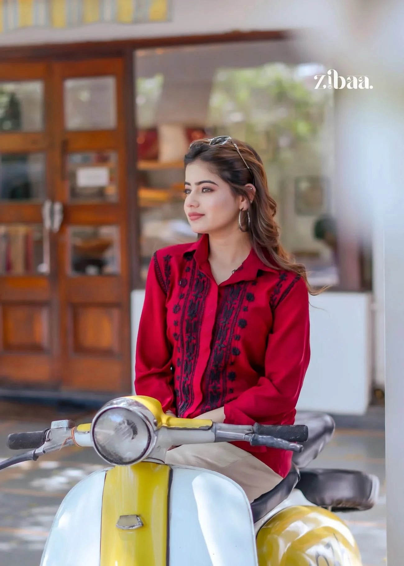 The model poses relaxed and casual while seated on the scooter, highlighting the fine black threadwork on her red chikankari shirt.