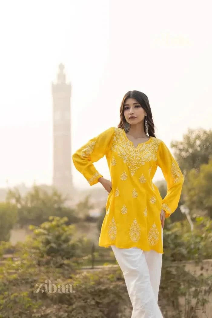 A woman wearing a yellow short kurti with intricate floral embroidery poses outdoors, hands on her hips, with a tower in the background.