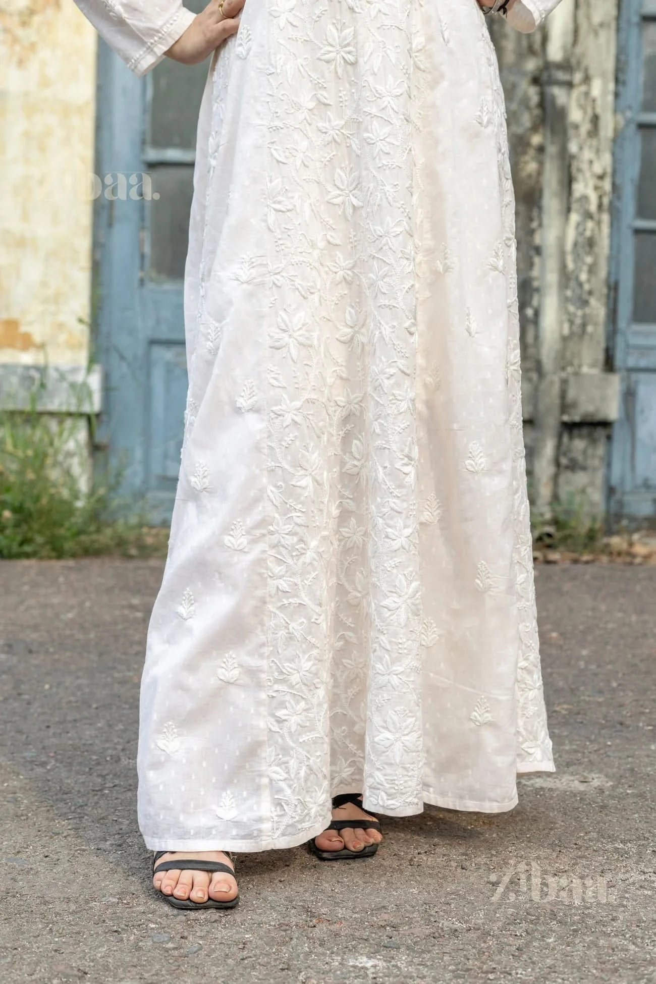 Close-up of a white Chikankari kurti with floral embroidery as the woman stands outdoors, hands resting on her hips, near a rustic door.