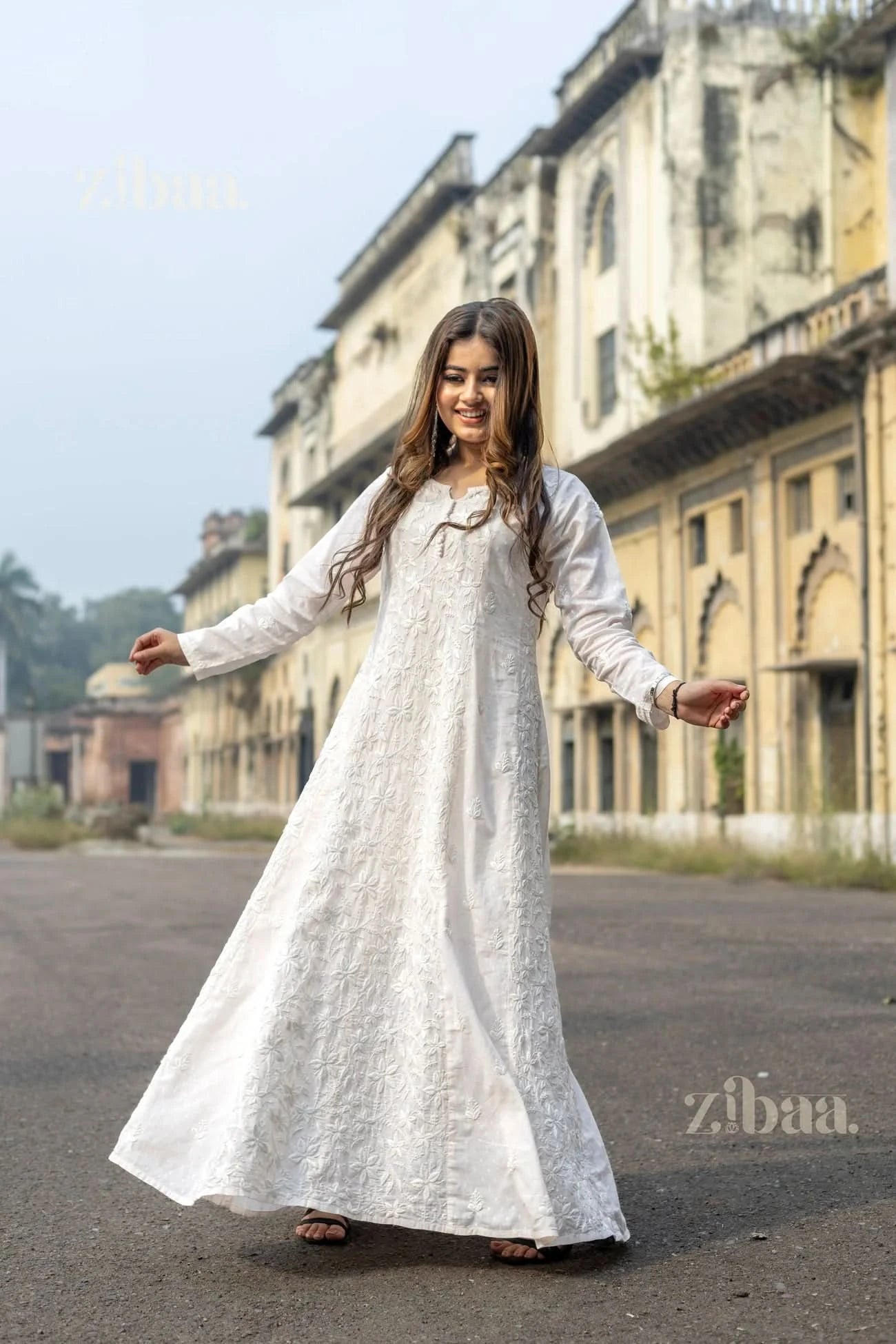 A woman wearing a white Chikankari kurti for women smiles brightly while twirling gracefully on an empty street near a rustic building.