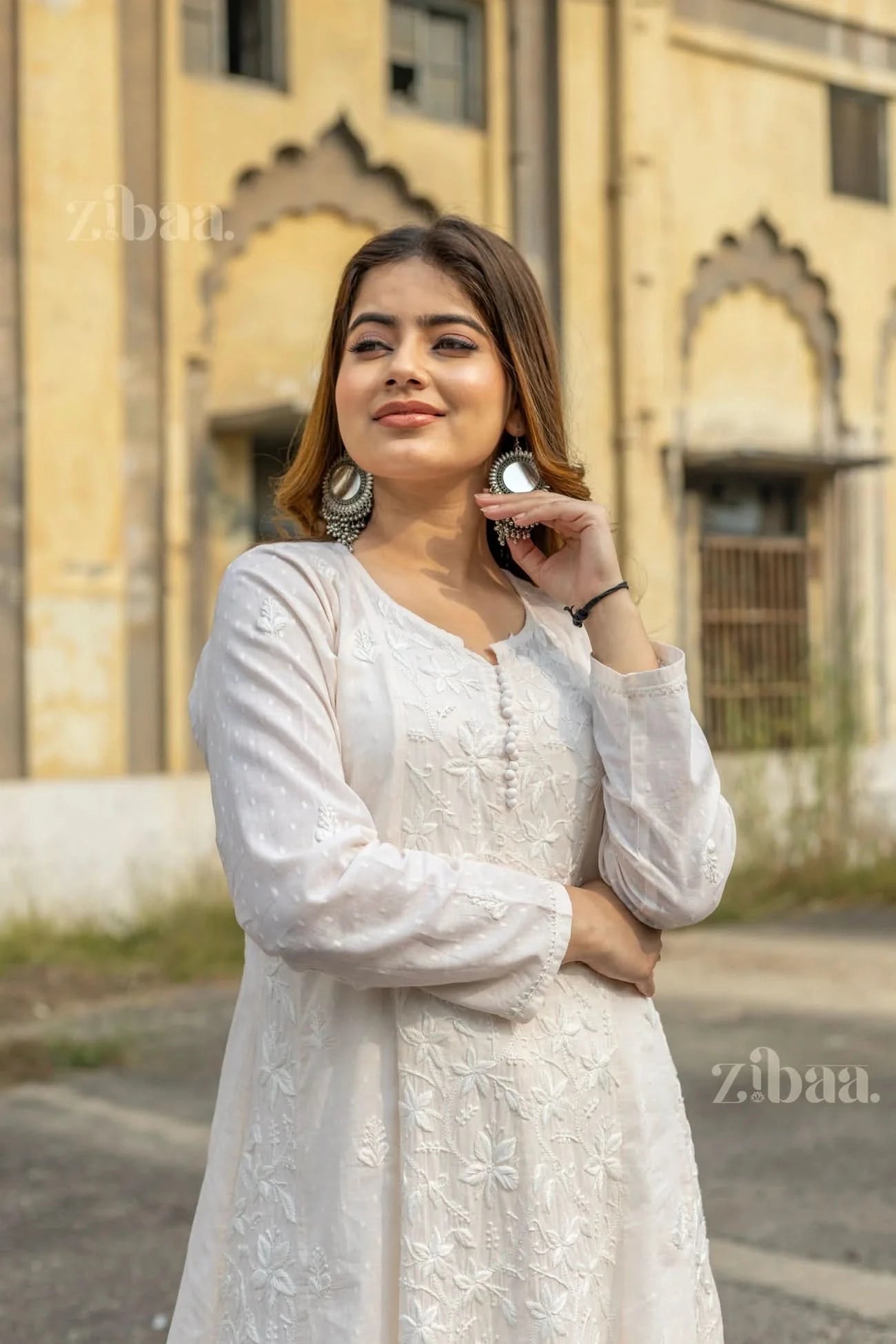 A woman wearing a white Chikankari kurti for women stands outdoors with her arms crossed, tilting her head slightly and smiling softly.