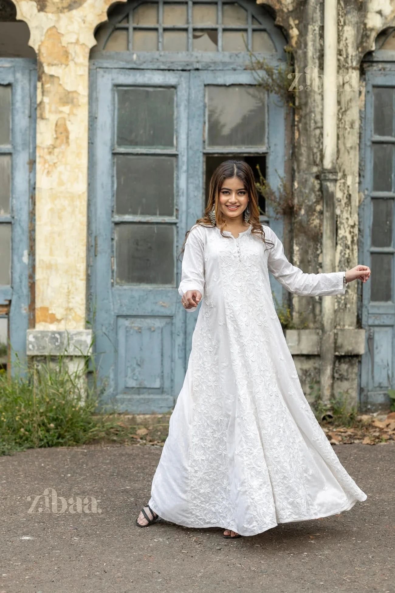 A woman wearing a flowy white Chikankari kurti with intricate embroidery smiles while twirling outdoors in front of a rustic blue door.