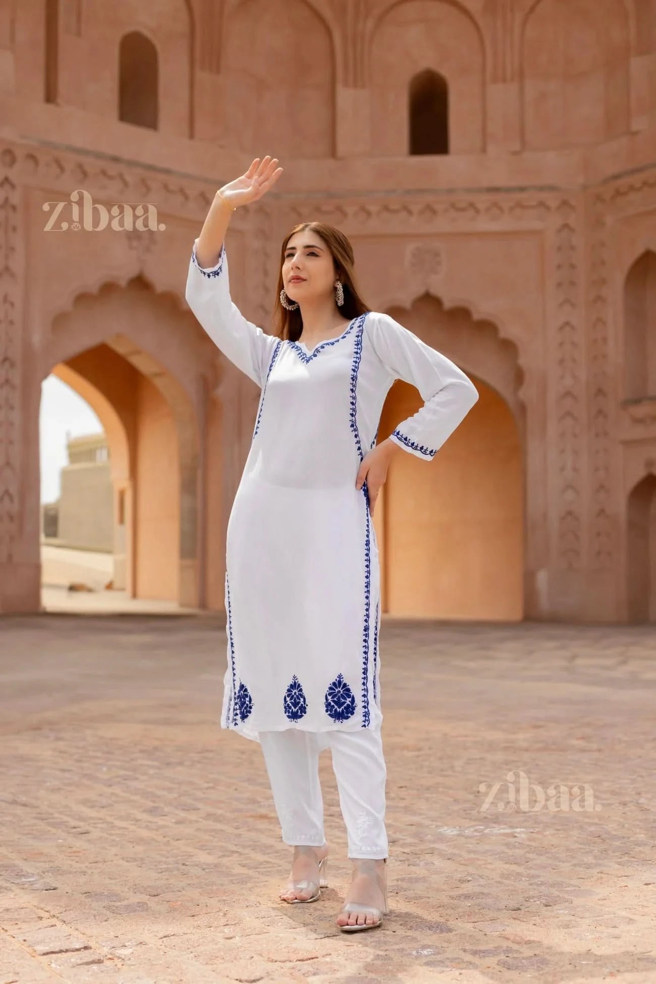 Woman in a white traditional outfit with blue patterns standing in front of architectural arches.