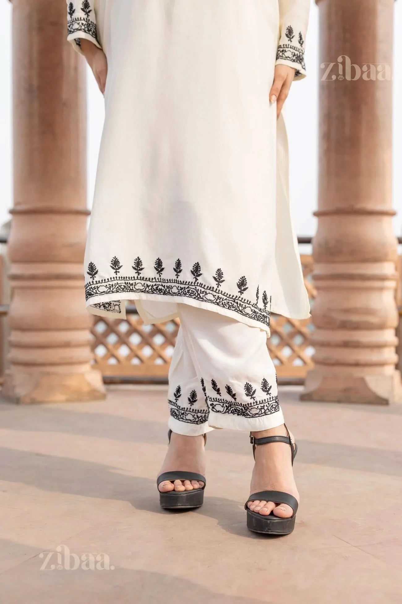 Close-up of a white kurta set for women with black embroidered details, as the woman stands in heels, hands resting by her sides.