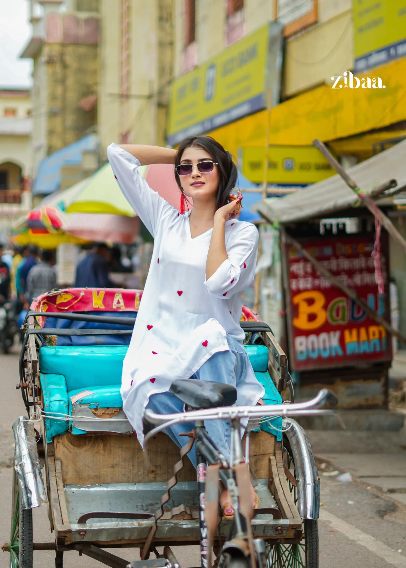 The model poses casually on a rickshaw, wearing a white chikankari kurti, with a bustling street and multiple shops visible in the background.