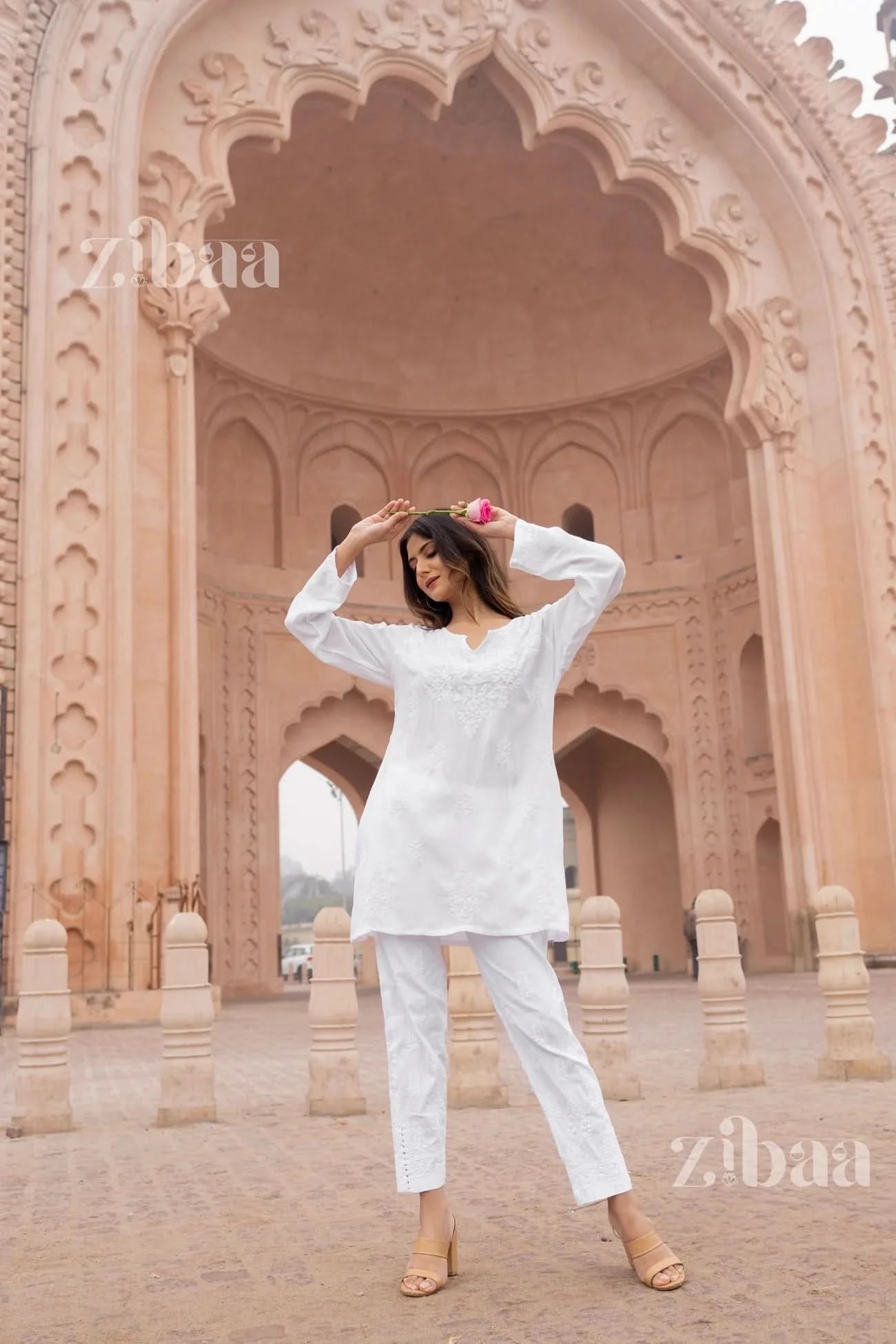A woman posing in a white short chikankari kurta with floral embroidery, round neck, and three-quarter sleeves, standing against an architectural backdrop.