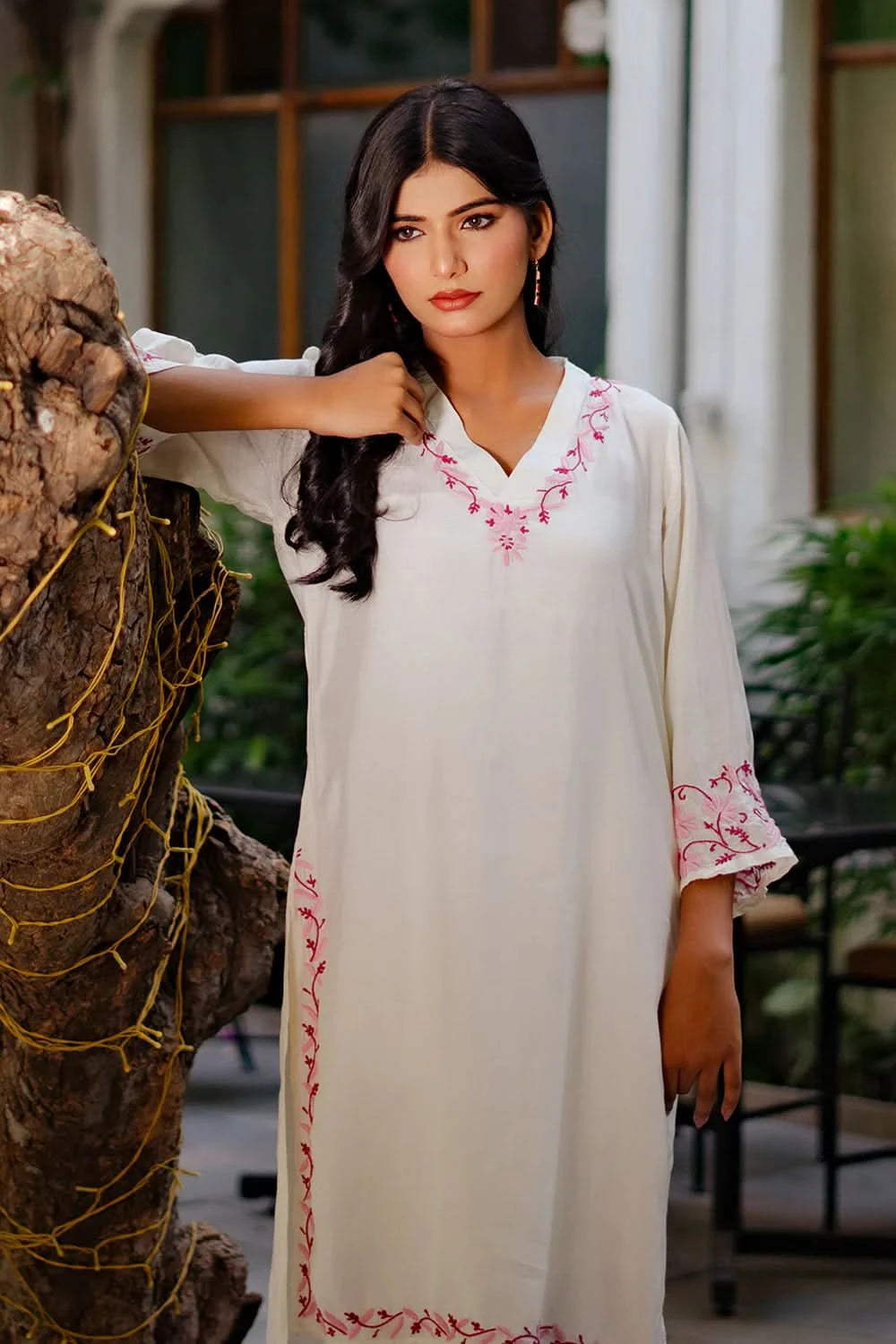 Woman wearing a white chikankari kurti with pink embroidery standing next to a textured wall.Woman wearing a white chikankari kurti with pink embroidery standing next to a textured wall.