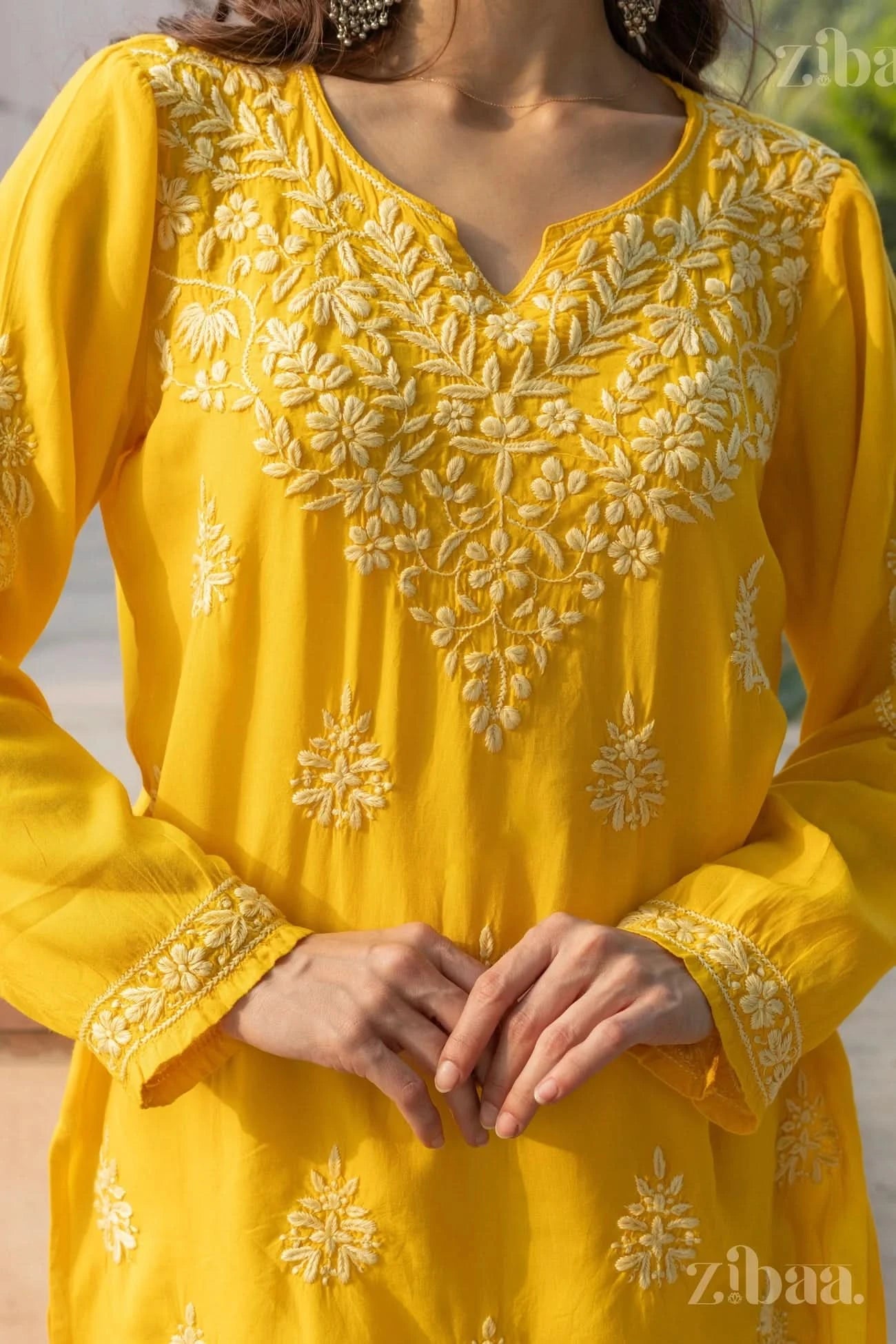 Close-up of a woman wearing a yellow Chikankari kurti with floral embroidery, standing outdoors with her hands gently clasped in front.