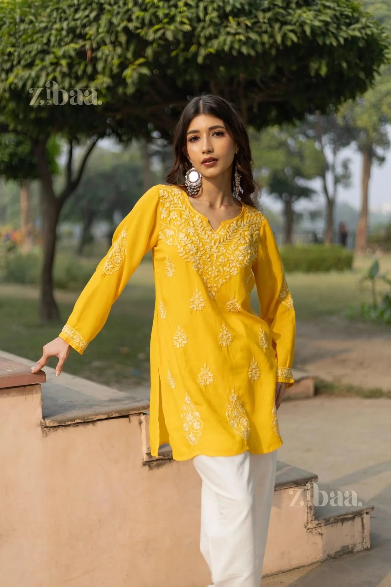A woman in a yellow short kurti with floral Chikankari embroidery poses outdoors, resting one hand on a ledge, looking confident.