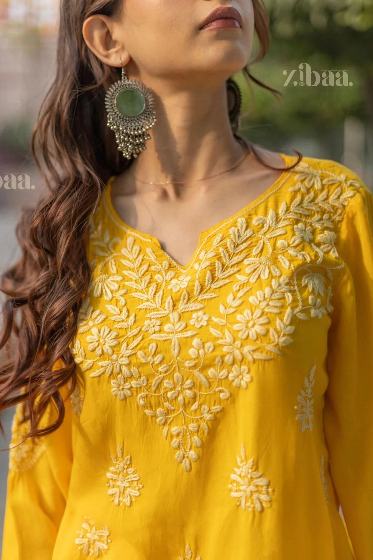 A woman wearing a yellow Chikankari kurti with floral embroidery stands outdoors, tilting her head slightly, showing her earrings.