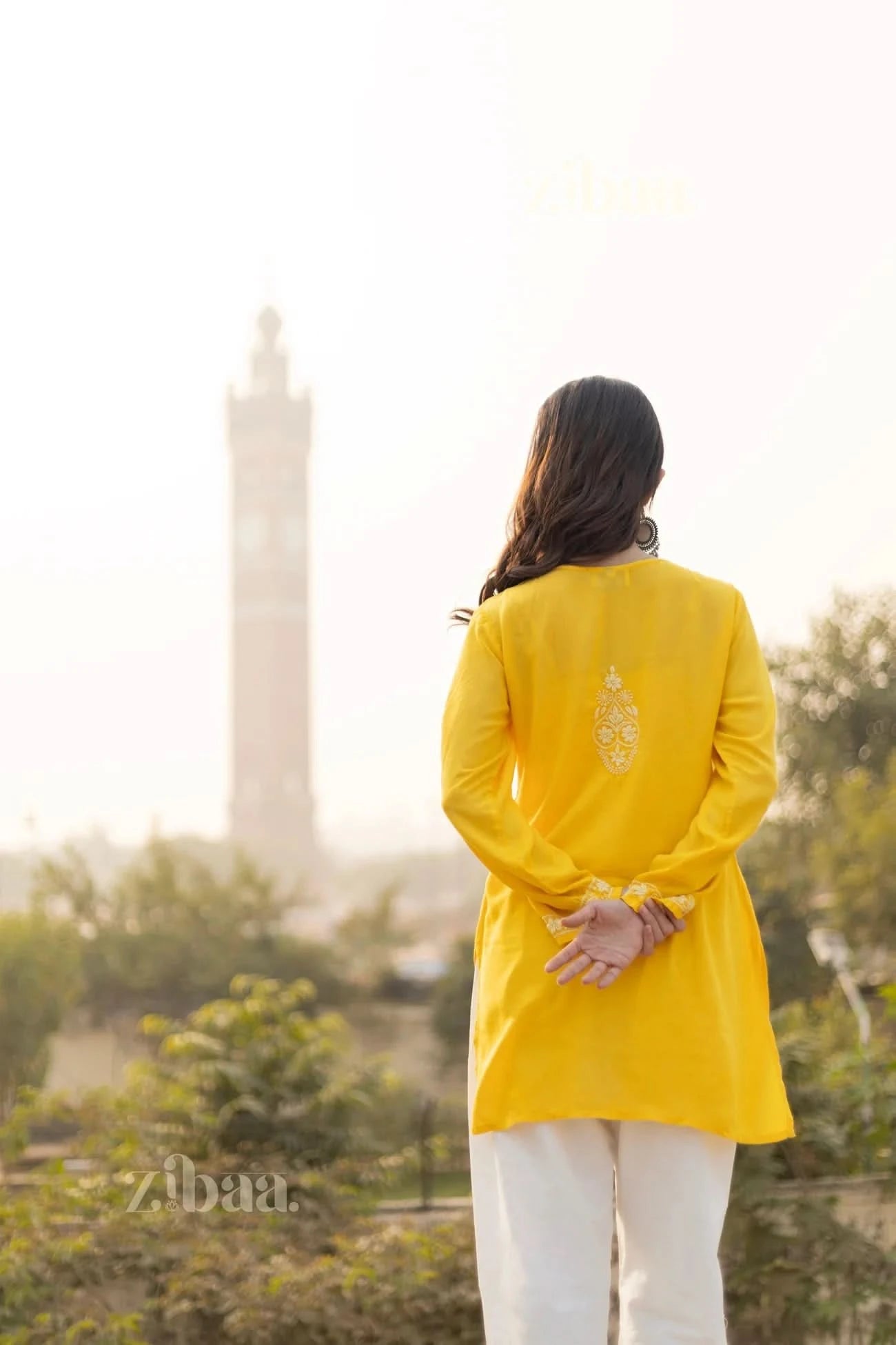 A woman in a yellow short kurti with back embroidery stands outdoors with her hands clasped behind her, facing a distant tower.