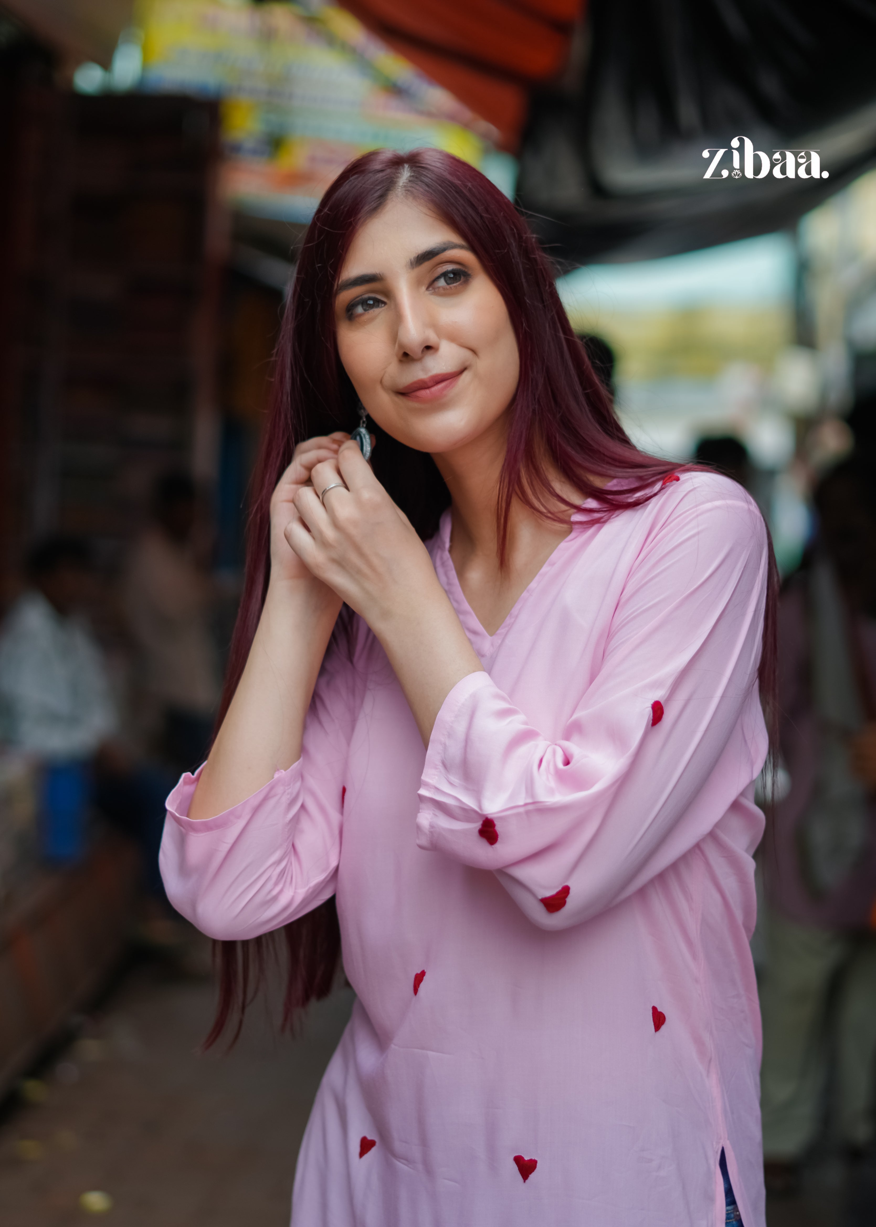 The woman in the  pink embroidered  hearts kurta, gently holding her hands near her face while posing with a soft smile.