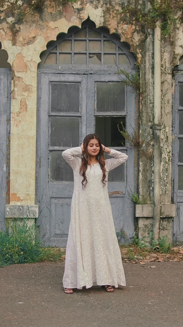 A woman wearing a white Chikankari Anarkali with intricate embroidery poses elegantly outside a heritage building in natural light.
