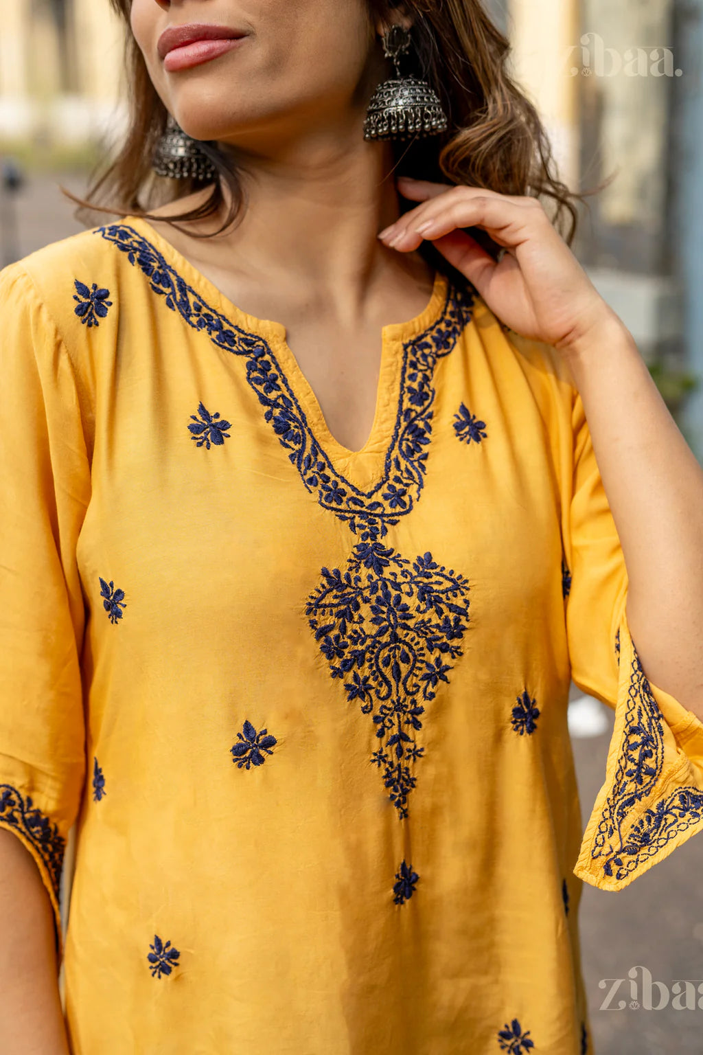 A girl poses outdoors in an eye catching mustard kurta with navy embroideries on the back and sleeves, with a pair of statement earrings.