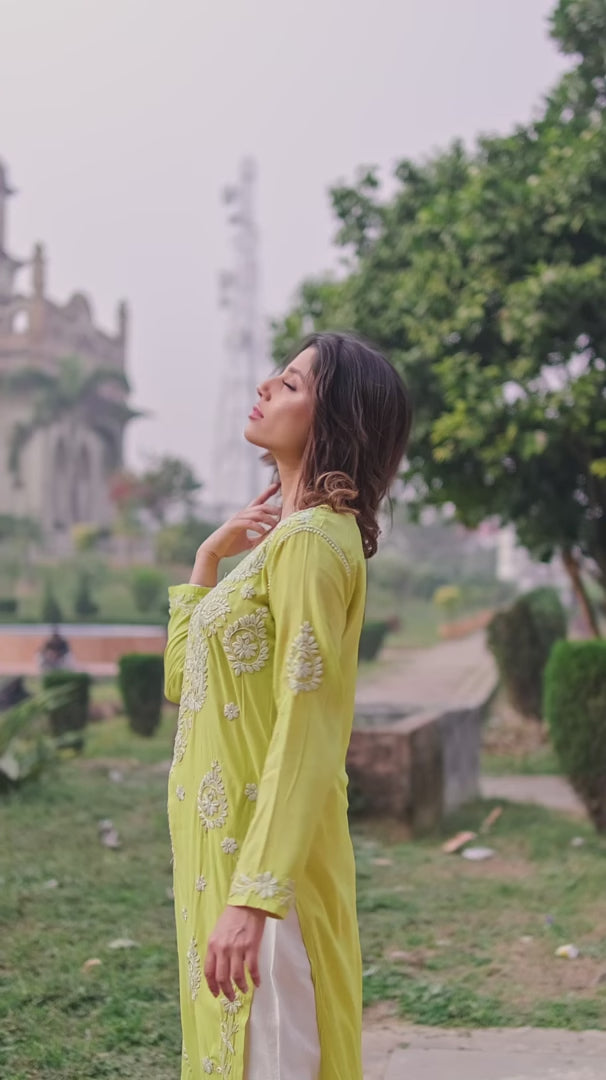 A woman leans against a textured wall in a lime green Chikankari Kurti with white embroidery, adjusting her hair, paired with white pants.