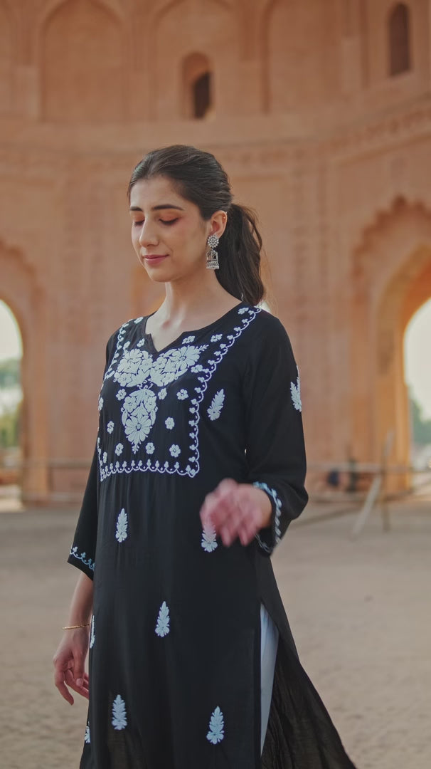 A girl in a black Chikankari kurti with intricate embroidery and transparent block-heeled sandals, posing in a heritage backdrop.